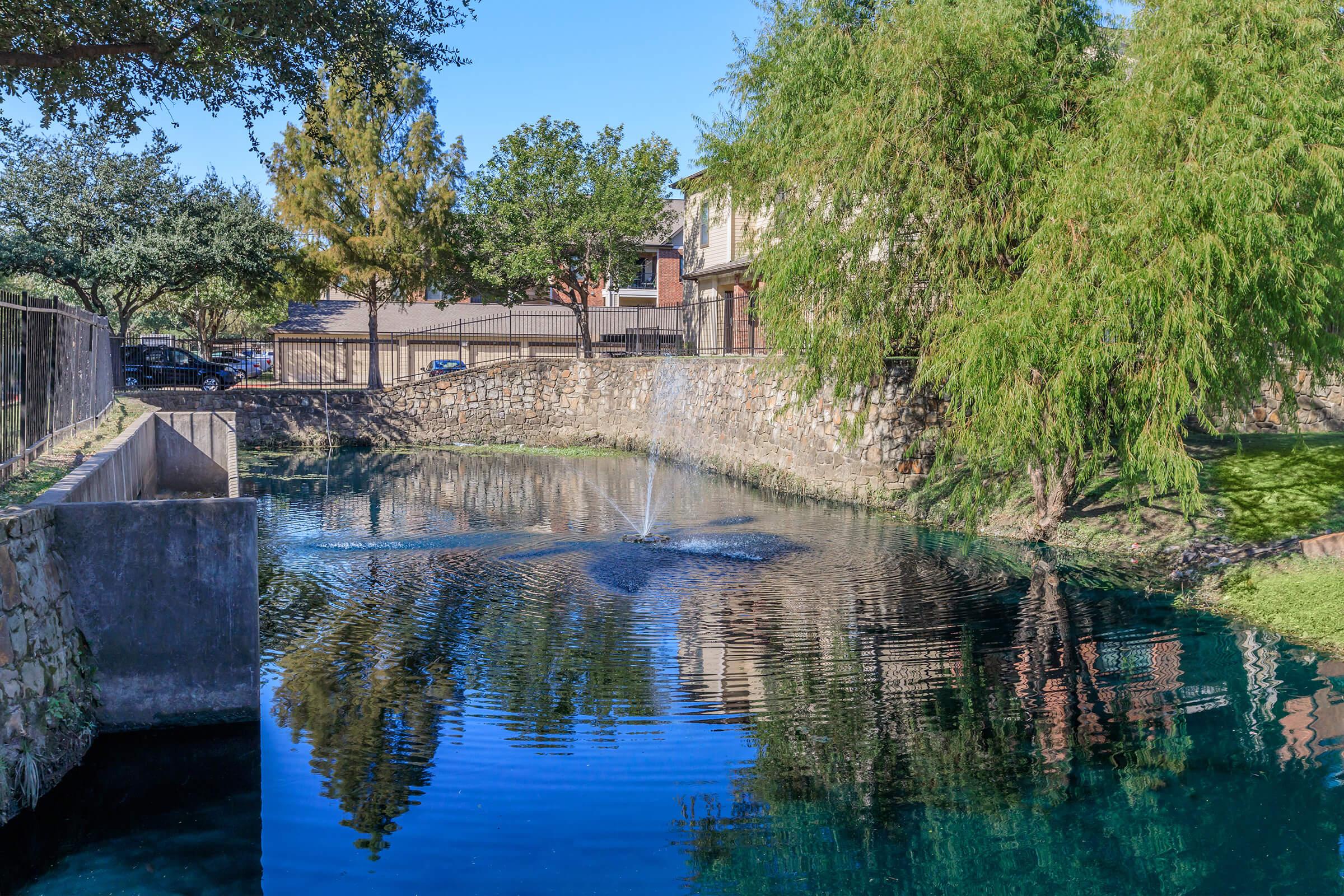 A serene pond surrounded by trees and stone walls. In the center, a small fountain creates ripples in the clear blue water. Residential buildings are visible in the background, and the sky is bright and clear, reflecting on the surface of the pond. The scene conveys a peaceful and well-maintained outdoor environment.