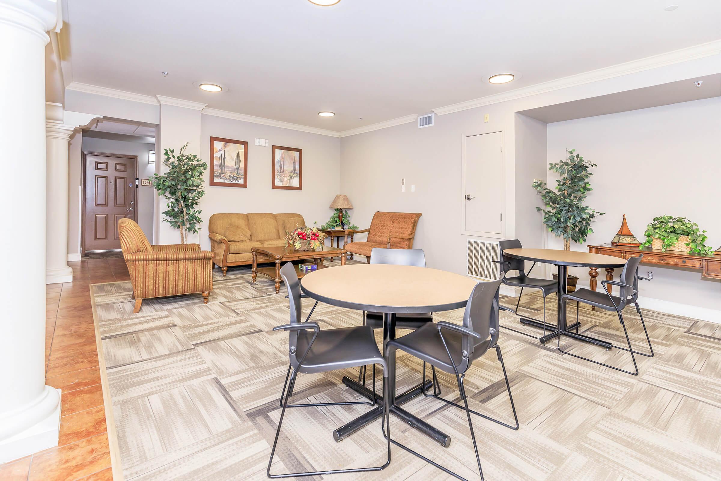 A cozy lounge area featuring comfortable seating, including a sofa and wicker chairs, hardwood floors, and decorative plants. There are two round tables with black chairs, framed artwork on the walls, and soft lighting creating a welcoming atmosphere.