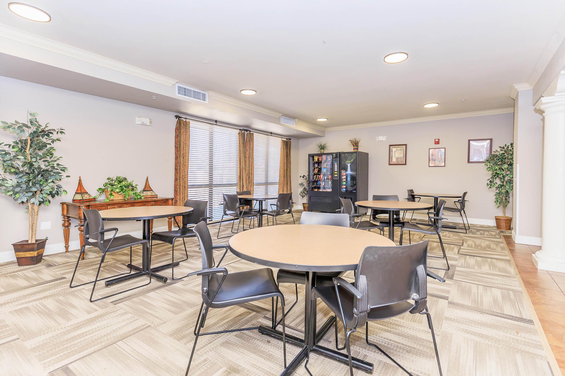 A spacious communal area featuring several round tables with black chairs, indoor plants, and windows with blinds. A snack vending machine stands in one corner, and framed art is displayed on the walls, creating a welcoming environment for social gatherings or leisure activities.