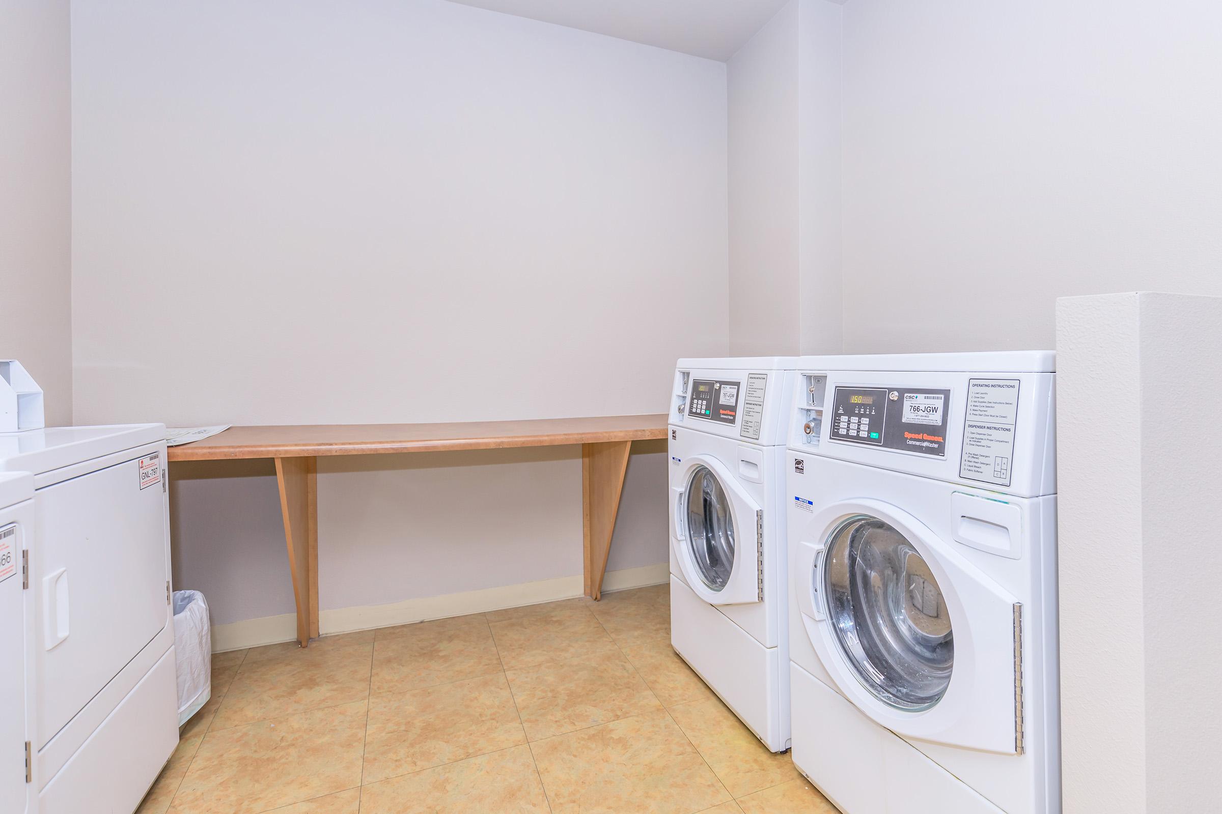 A clean laundry room featuring two stacked washing machines and dryers against a light-colored wall. A wooden countertop is available for folding laundry, and there is a wastebasket nearby. The floor is tiled in a neutral color, creating a functional and organized space.