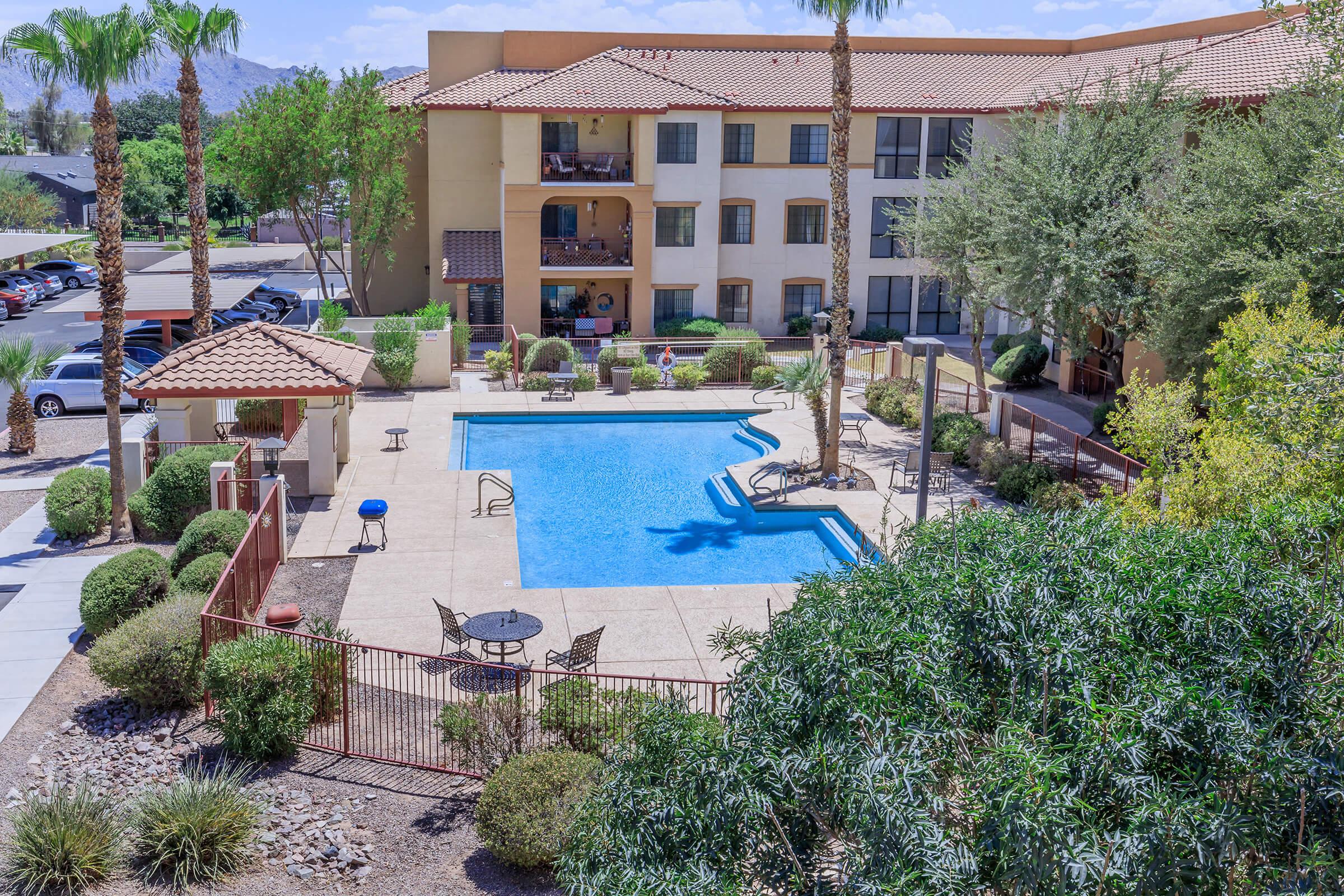 Aerial view of a hotel pool area surrounded by palm trees and shrubs. The rectangular blue pool is fenced, with lounge chairs and tables nearby. The hotel has several floors and balconies overlooking the pool. Bright, sunny day with clear skies and mountainous background.