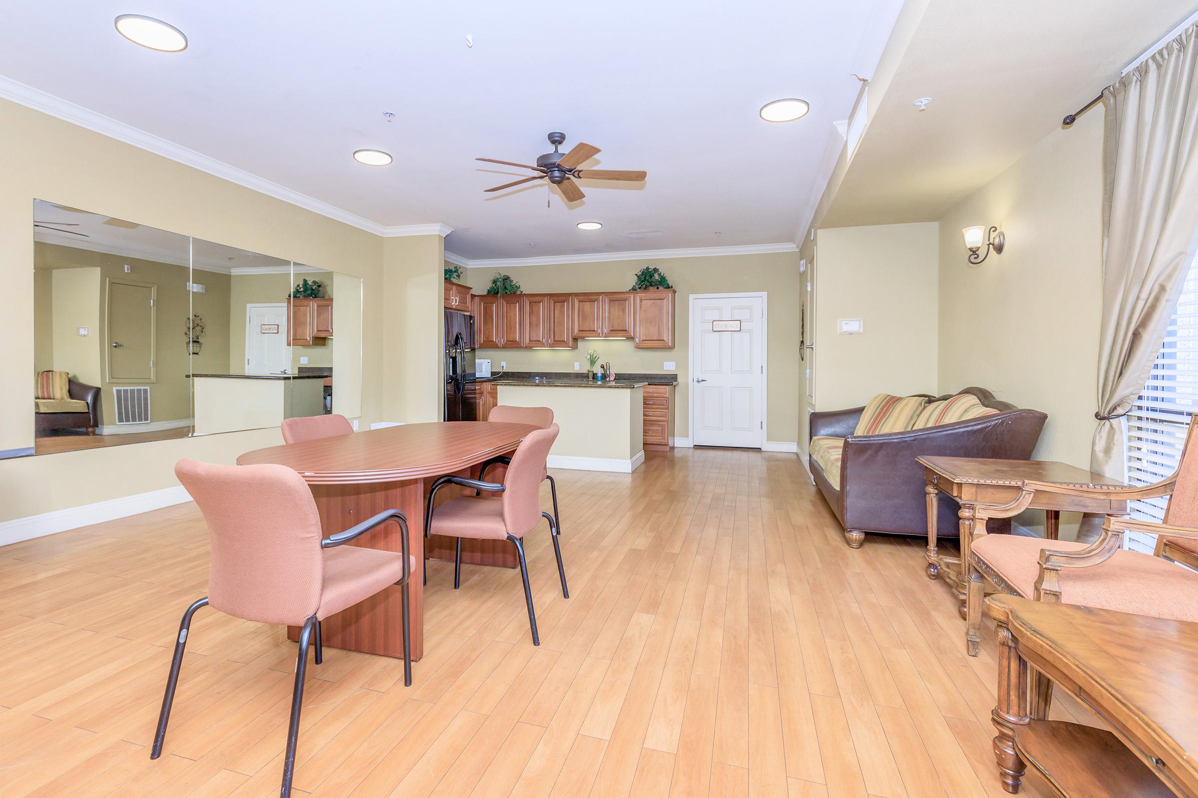 Interior view of a modern living space featuring a dining table with chairs, a kitchen area in the background, and a comfortable seating arrangement including a sofa. The room has light-colored walls, wooden flooring, and decorative plants. Natural light comes through the windows, creating a warm atmosphere.