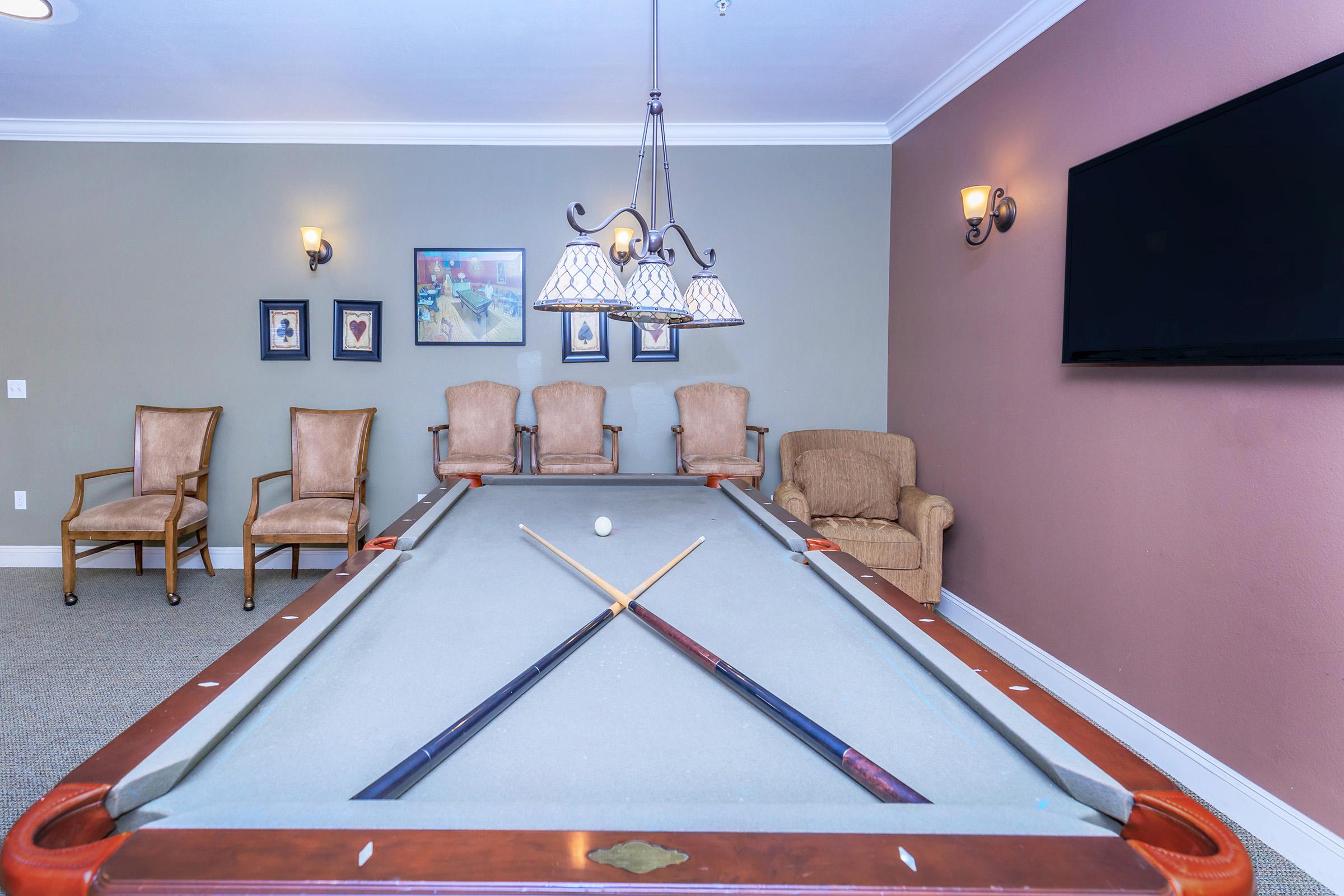 A billiards table with crossed cues and a single white ball in the center. In the background, there are four upholstered chairs and a cozy armchair, along with framed photos on the wall. A chandelier hangs above the table, and a flat-screen TV is mounted on the wall to the right.