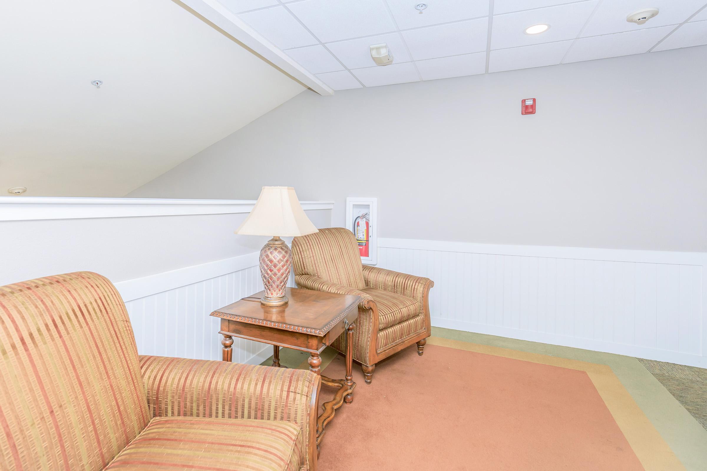 A cozy seating area featuring two striped armchairs flanking a wooden side table with a lamp. The room has light-colored walls and a simple rug, creating a welcoming atmosphere.