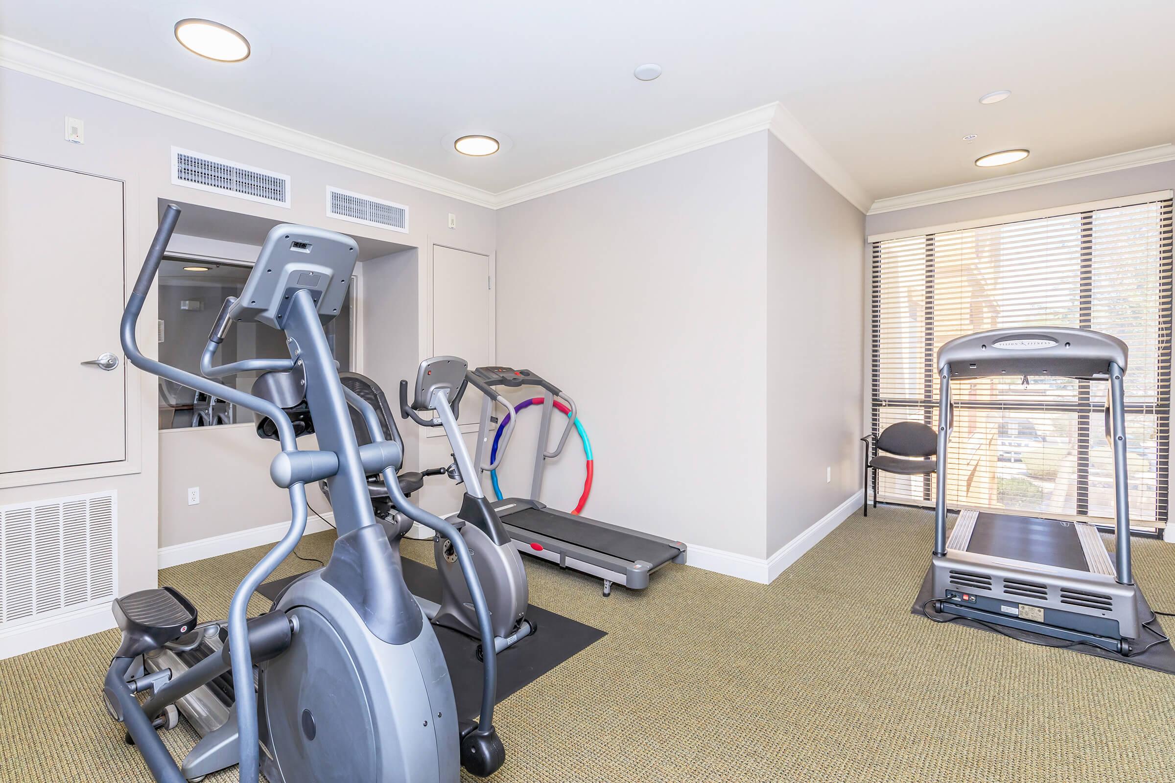 Interior of a gym featuring various exercise equipment, including a treadmill, elliptical machine, and a stationary bike. The space has neutral wall colors, ample natural light from large windows, and a carpeted floor. There are also colorful exercise hoops in the corner and a chair against the wall.