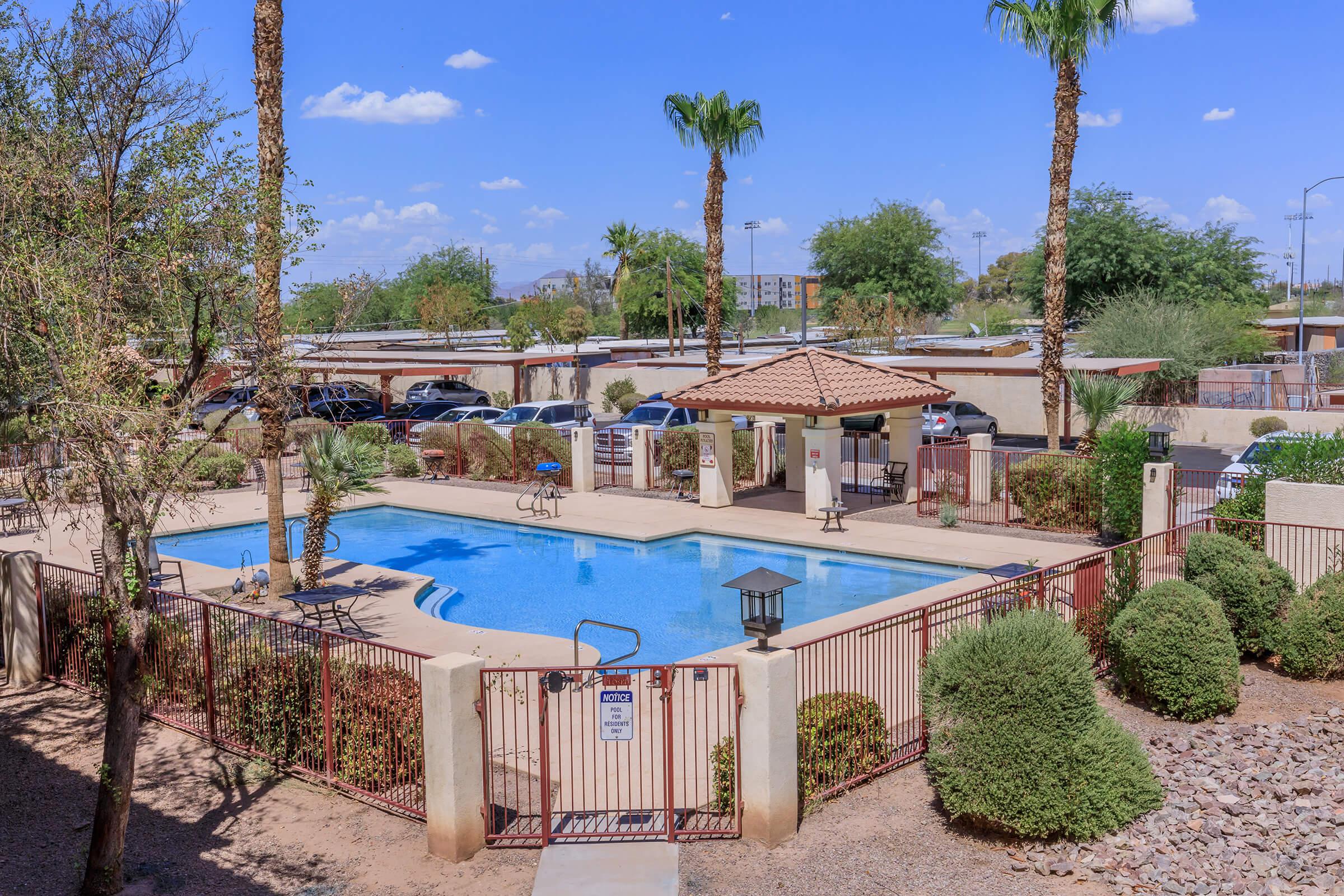 A clear blue swimming pool surrounded by palm trees and landscaped greenery. There are lounge chairs around the pool, and a covered gazebo nearby. In the background, parked cars are visible along a paved area, with the sky showing a few fluffy clouds.
