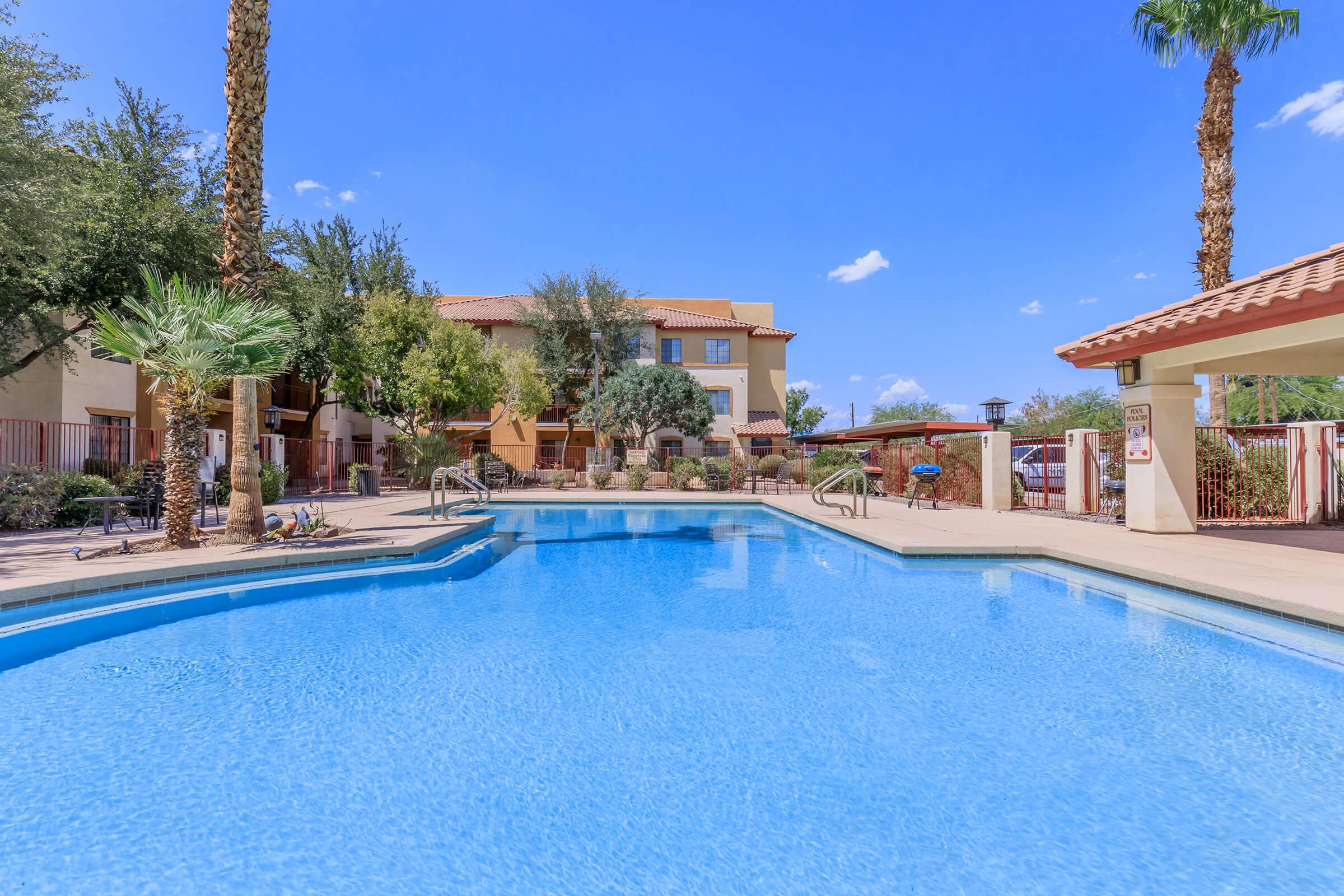 A clear blue swimming pool with shallow steps leads into the water. Lush palm trees and landscaped greenery surround the pool area, which is set against a backdrop of a multi-story building. The sky is bright blue with a few clouds. Sunlight creates a vibrant and inviting atmosphere.