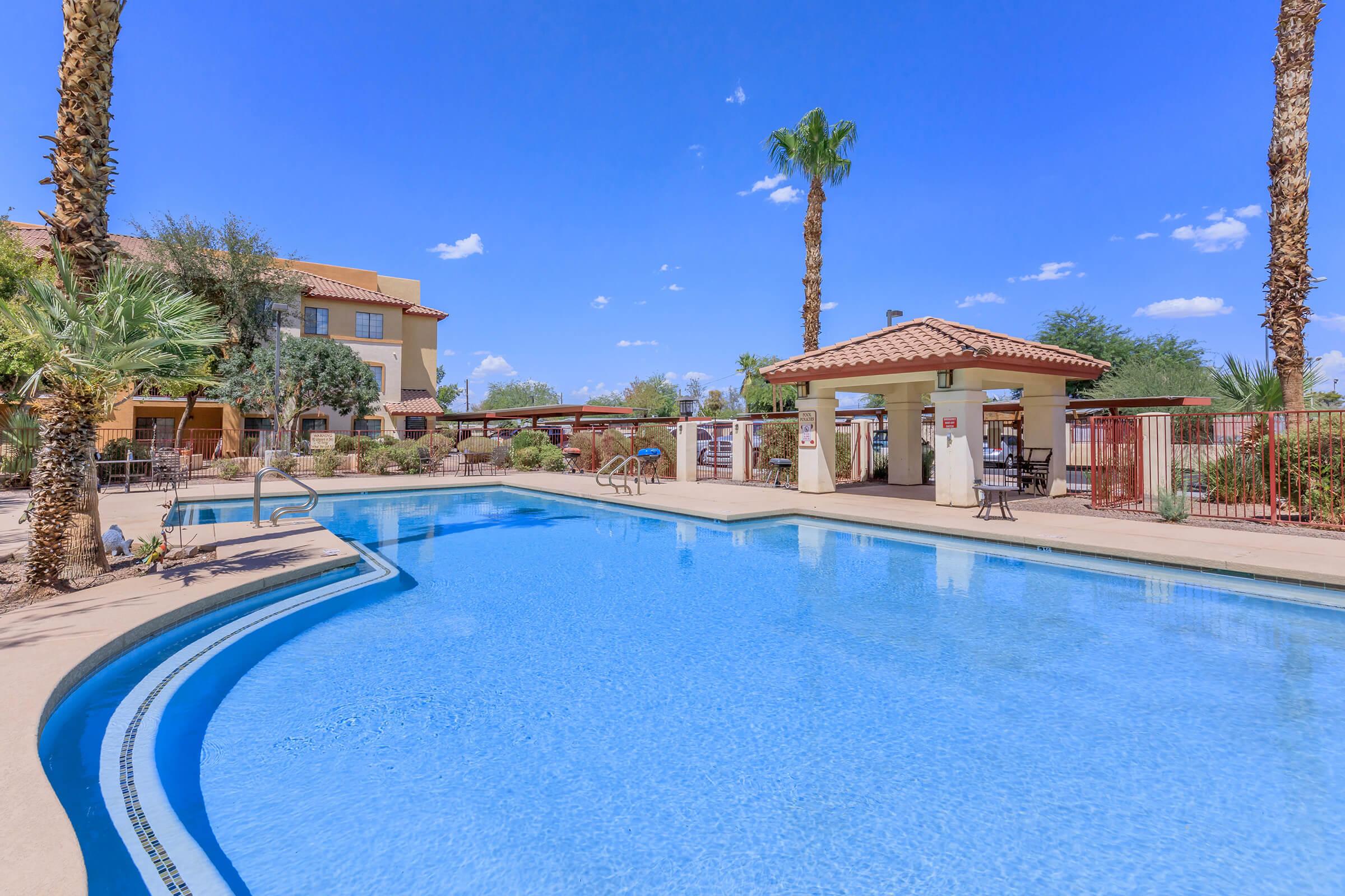 A sunny outdoor pool area featuring clear blue water, surrounded by palm trees and a well-maintained landscape. Lounge chairs are placed around the pool, and a shaded gazebo is visible nearby. The sky is bright blue with a few clouds, and a building is situated in the background.