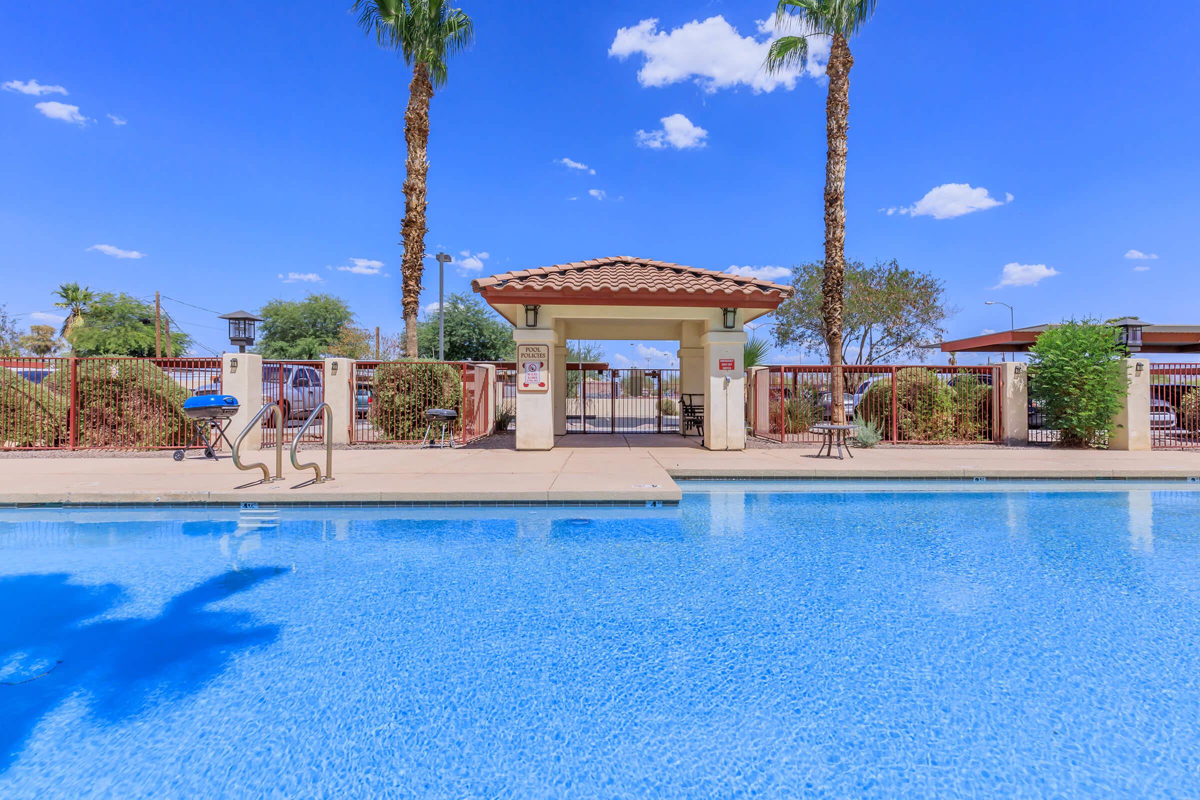 View of a sparkling blue swimming pool with a gated entrance, surrounded by palm trees and lush landscaping. The clear sky is bright with a few fluffy clouds. A set of stairs leads into the pool, and nearby seating is visible, creating an inviting atmosphere for relaxation.
