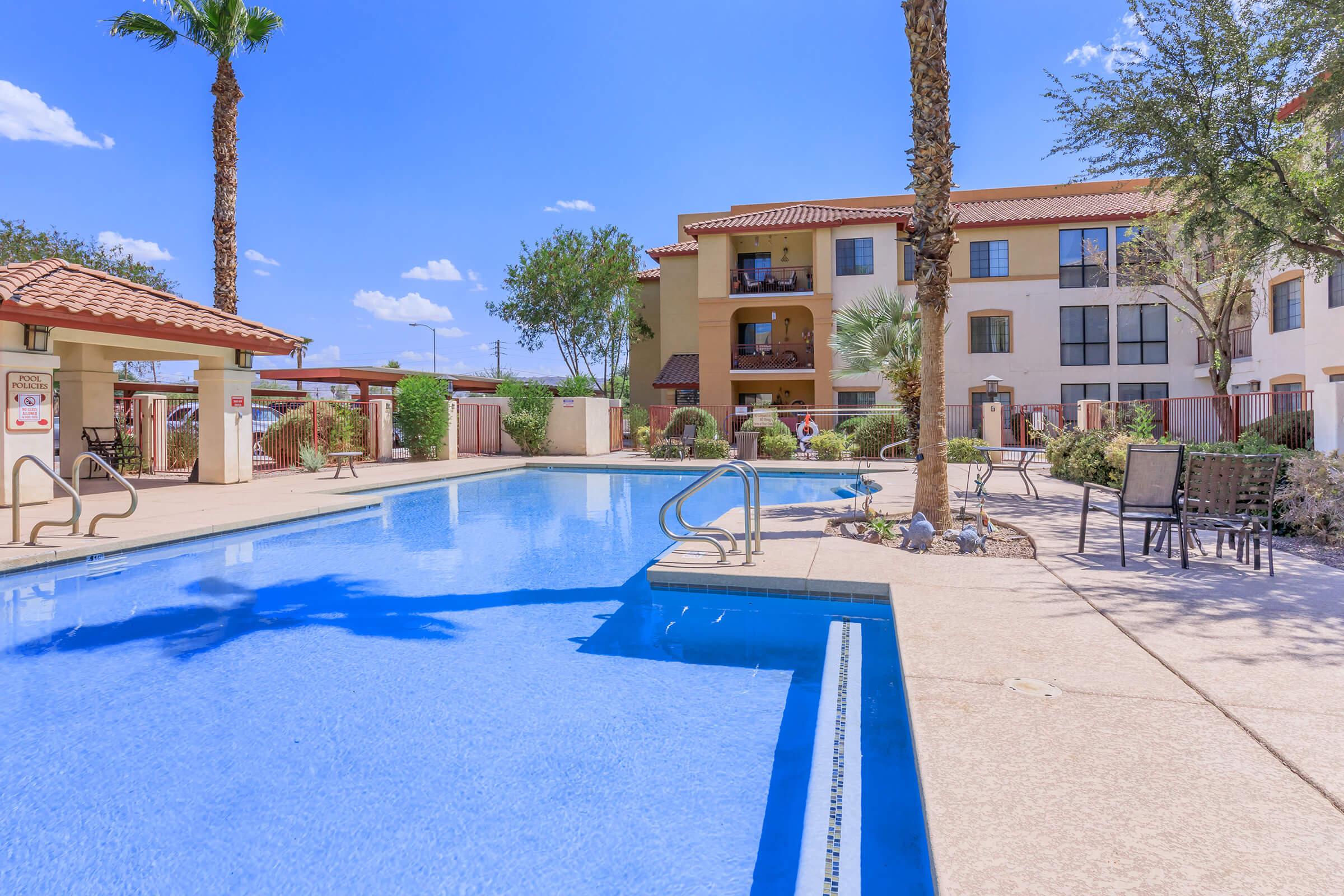 A clear blue swimming pool surrounded by palm trees and lounge chairs, with a multi-story building in the background. The sky is bright and sunny, with a few clouds. The scene evokes a relaxing outdoor atmosphere, ideal for leisure and recreation.