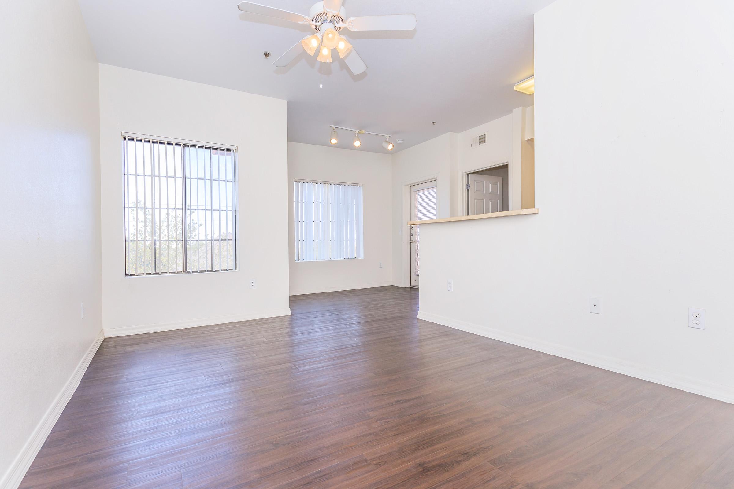 A spacious, empty room featuring light-colored walls and a wooden floor. Large windows with vertical blinds allow natural light to enter. A ceiling fan hangs from the ceiling, and there are track lights along the wall. The space has an open layout leading to another area, suggesting versatility for furniture arrangement.