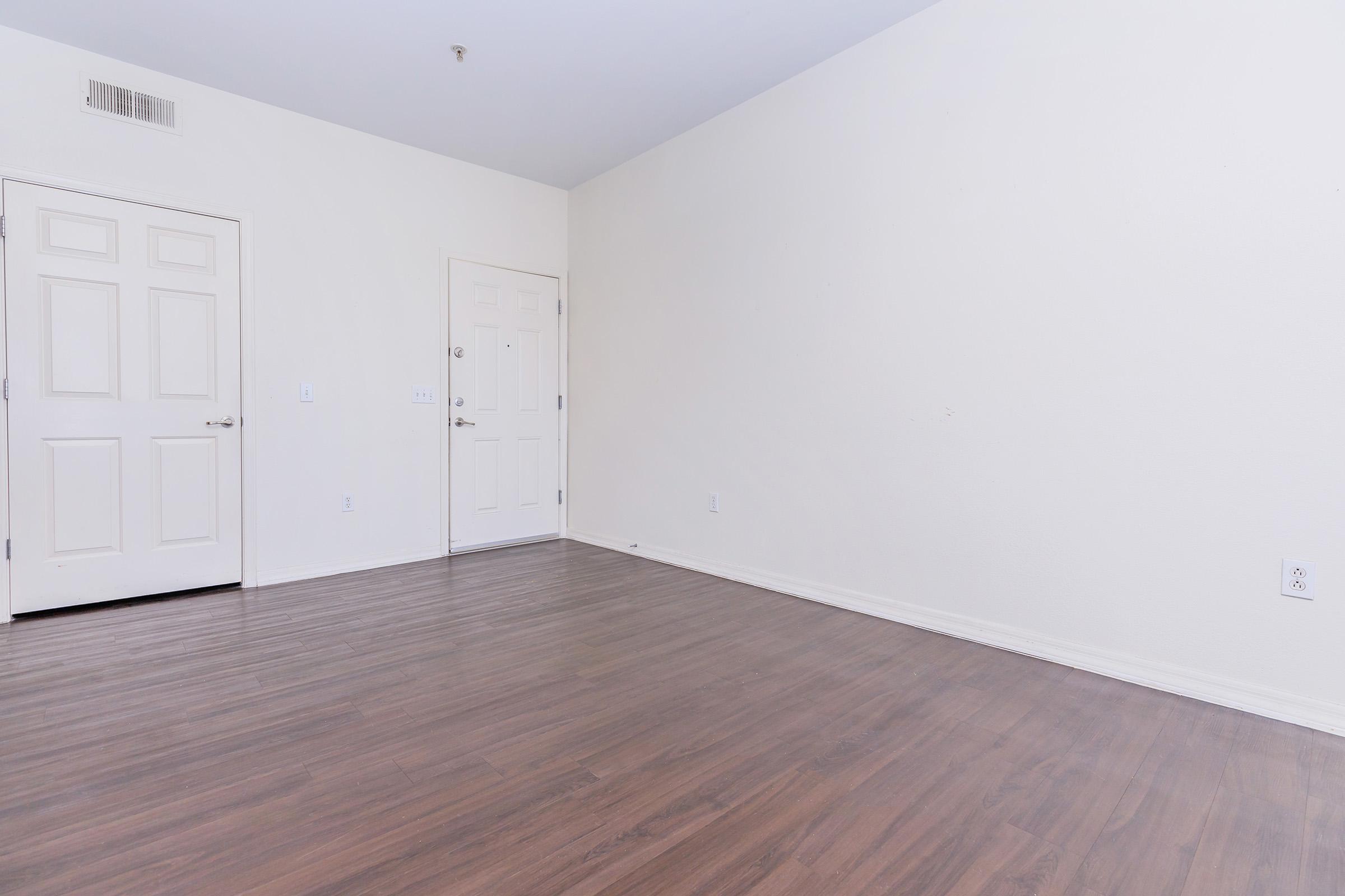 A minimalist, empty room featuring light-colored walls and a wooden floor. There are two closed doors; one is white with a simple design, and the other is a standard entry door. The space is well-lit and has no furniture, creating a bright and open atmosphere.