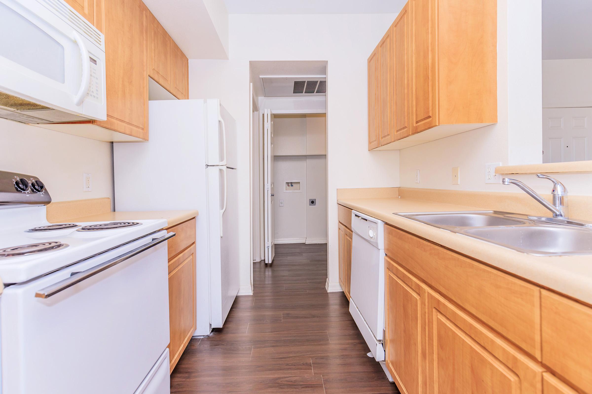 Modern kitchen with wooden cabinets and a light countertop. Features a white stove, microwave, and refrigerator, along with a double sink and dishwasher. The room has a clean, spacious layout and leads to a hallway with a closet. The floor is dark wood, creating a warm ambiance.