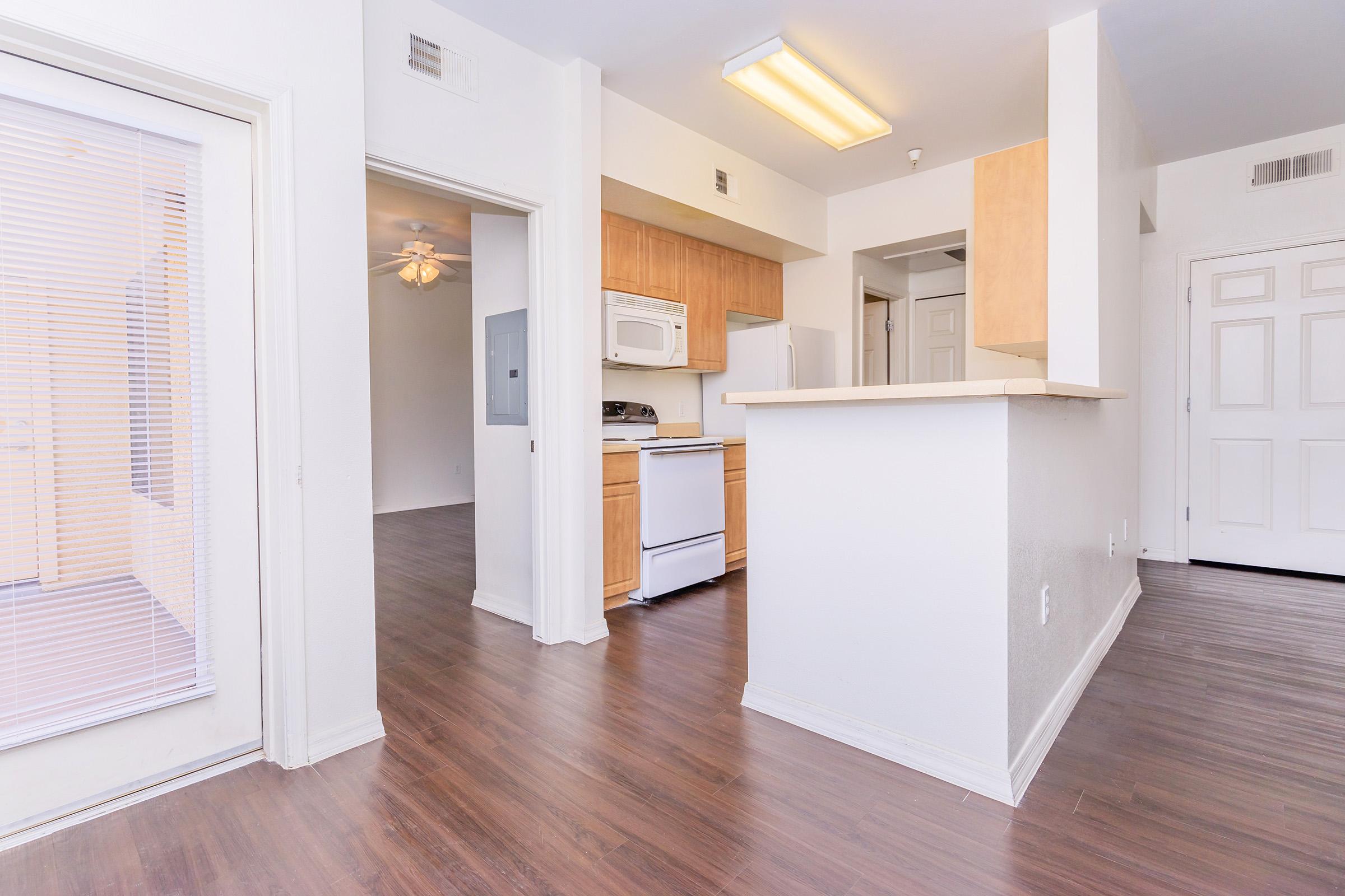 A bright, spacious apartment interior featuring a kitchen with wooden cabinets, a white refrigerator, and a stove. The room has laminate flooring and is well-lit with natural light coming from a nearby window. Doorways lead to other areas of the apartment, creating an open feel.