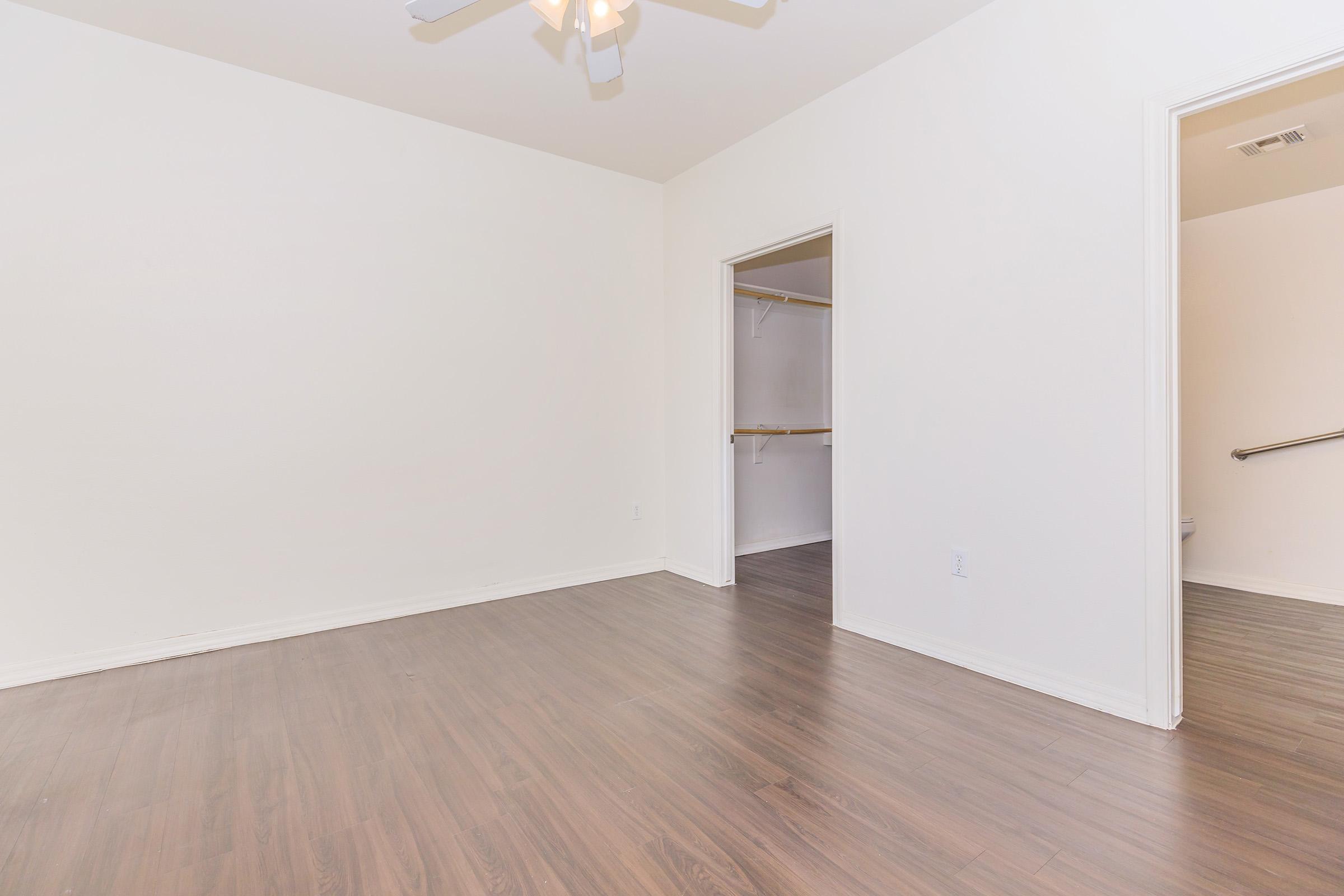 Empty room featuring light-colored walls, a ceiling fan, and brown laminate flooring. There are two doorways: one leading to a closet with shelving, and another that opens into a hallway or bathroom. The space is bright and uncluttered, creating a minimalistic environment.