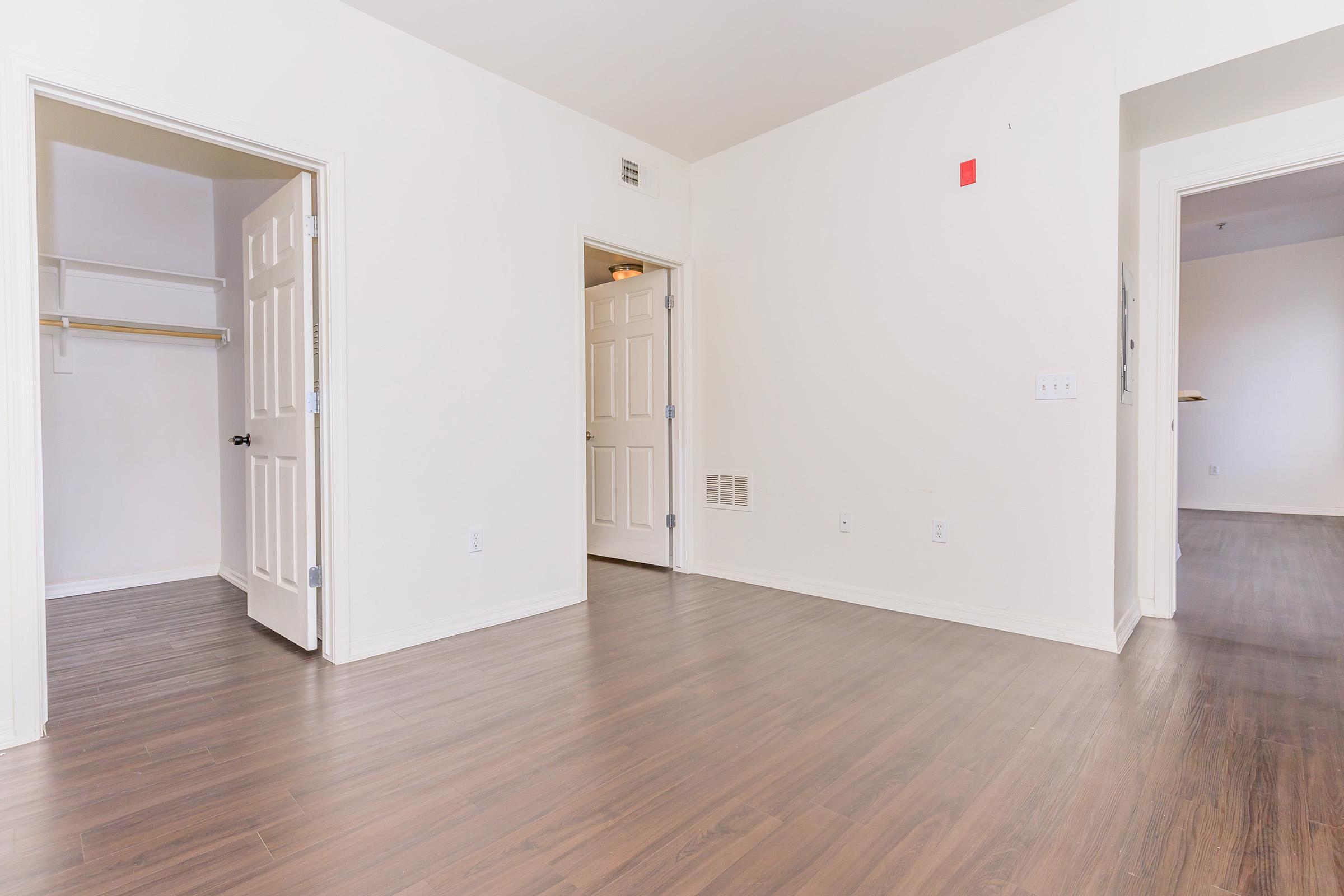 A spacious interior room featuring light-colored walls, smooth wooden flooring, and open doorways leading to adjacent rooms. One doorway opens to a closet with a shelf, while the other leads to an undefined space. The room has ample natural light and a modern feel.