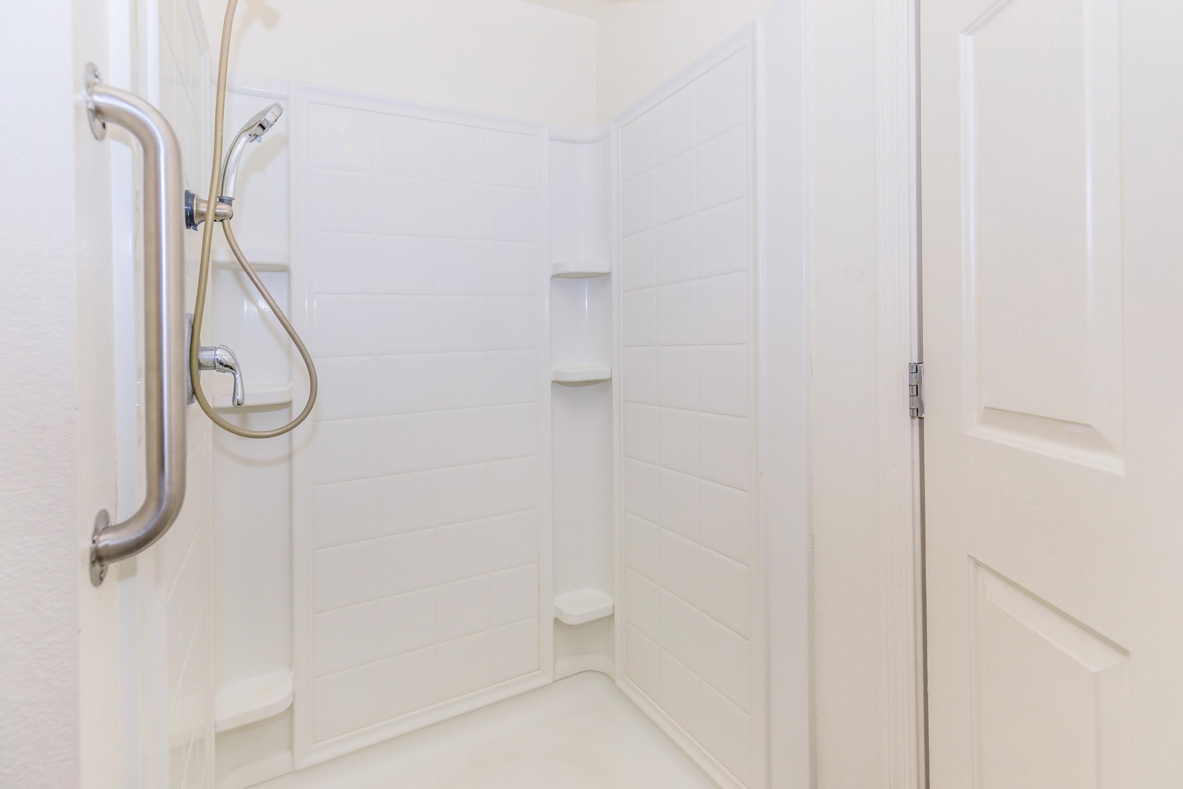 A clean, white shower stall with a handheld showerhead mounted on the wall. The stall features a textured floor, tiled walls, and a grab bar for safety. There are built-in shelves for toiletries, and a closed white door is visible to the right. The lighting is bright and the overall appearance is modern and minimalist.