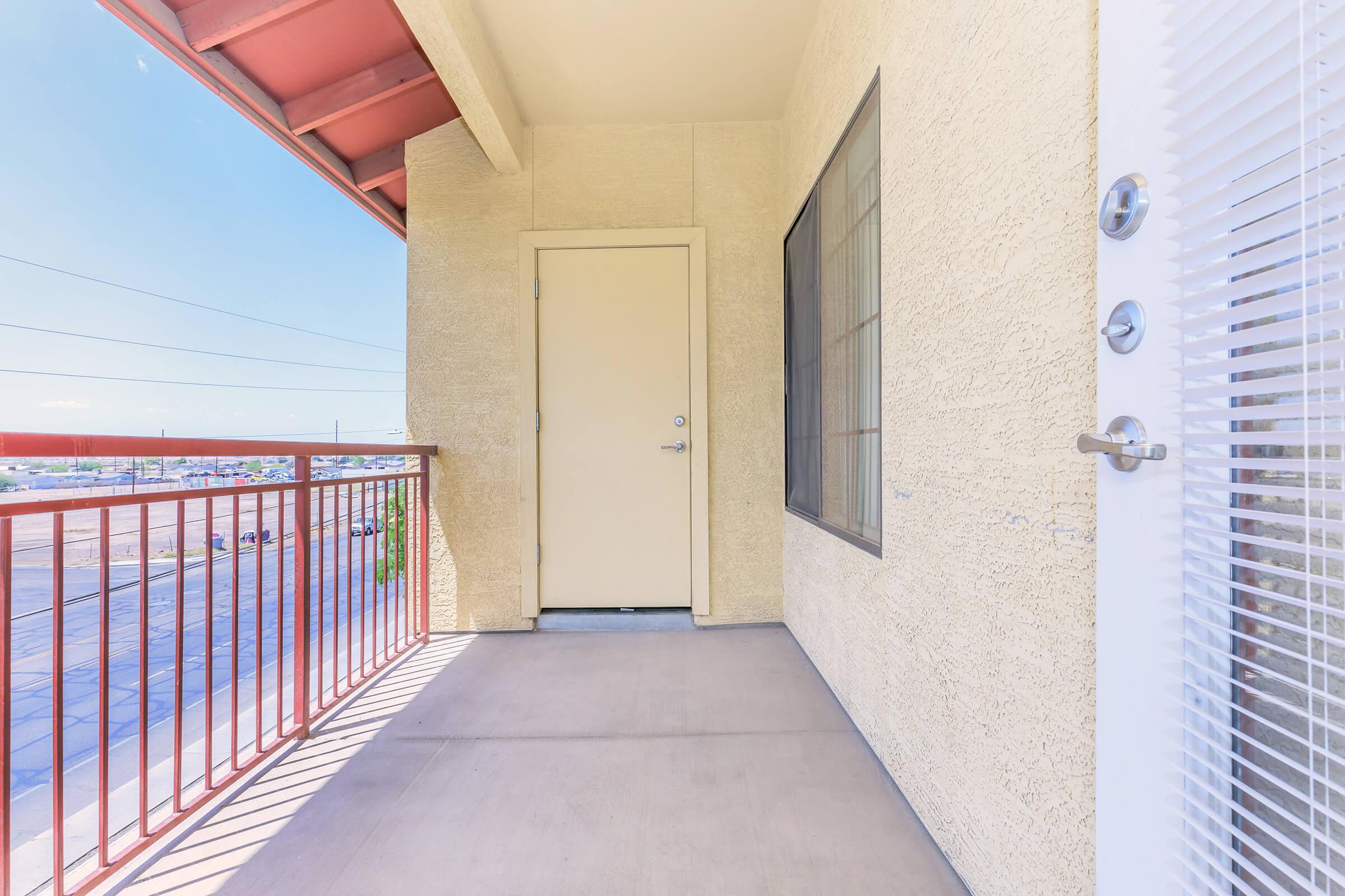 A view of a small balcony with a beige wall and a door leading to an interior space. The railing is red, creating a contrast with the light-colored wall. A window is visible next to the door, and the scene shows a clear blue sky in the background. The view includes distant buildings and power lines.