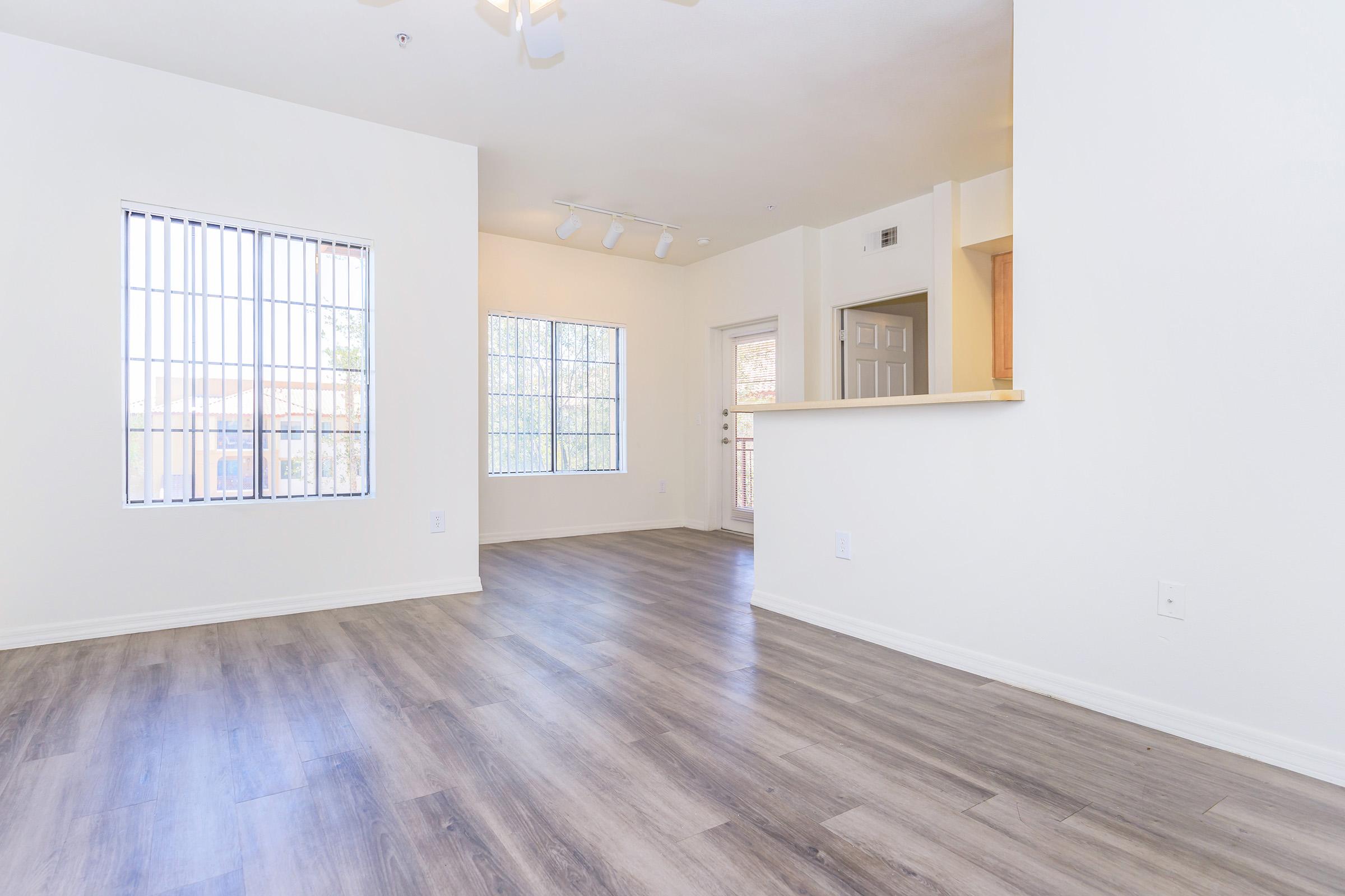 A spacious, empty room with light-colored walls and large windows featuring vertical blinds. The floor is covered with light wood-like laminate. There is a doorway leading to a kitchen area and another door that opens to an outside balcony. Natural light fills the space, creating an inviting atmosphere.