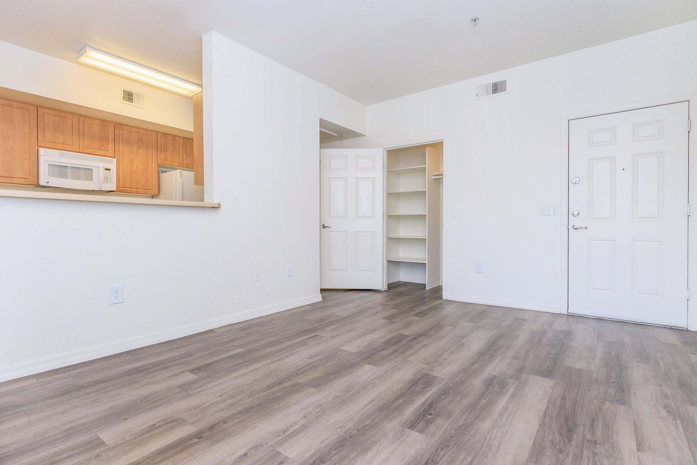 A spacious, empty room featuring light-colored walls and wooden flooring. The entrance is visible, along with a kitchen area in the background that has wooden cabinetry and a microwave. A closet with shelves is situated adjacent to the door, and natural light brightens the space.