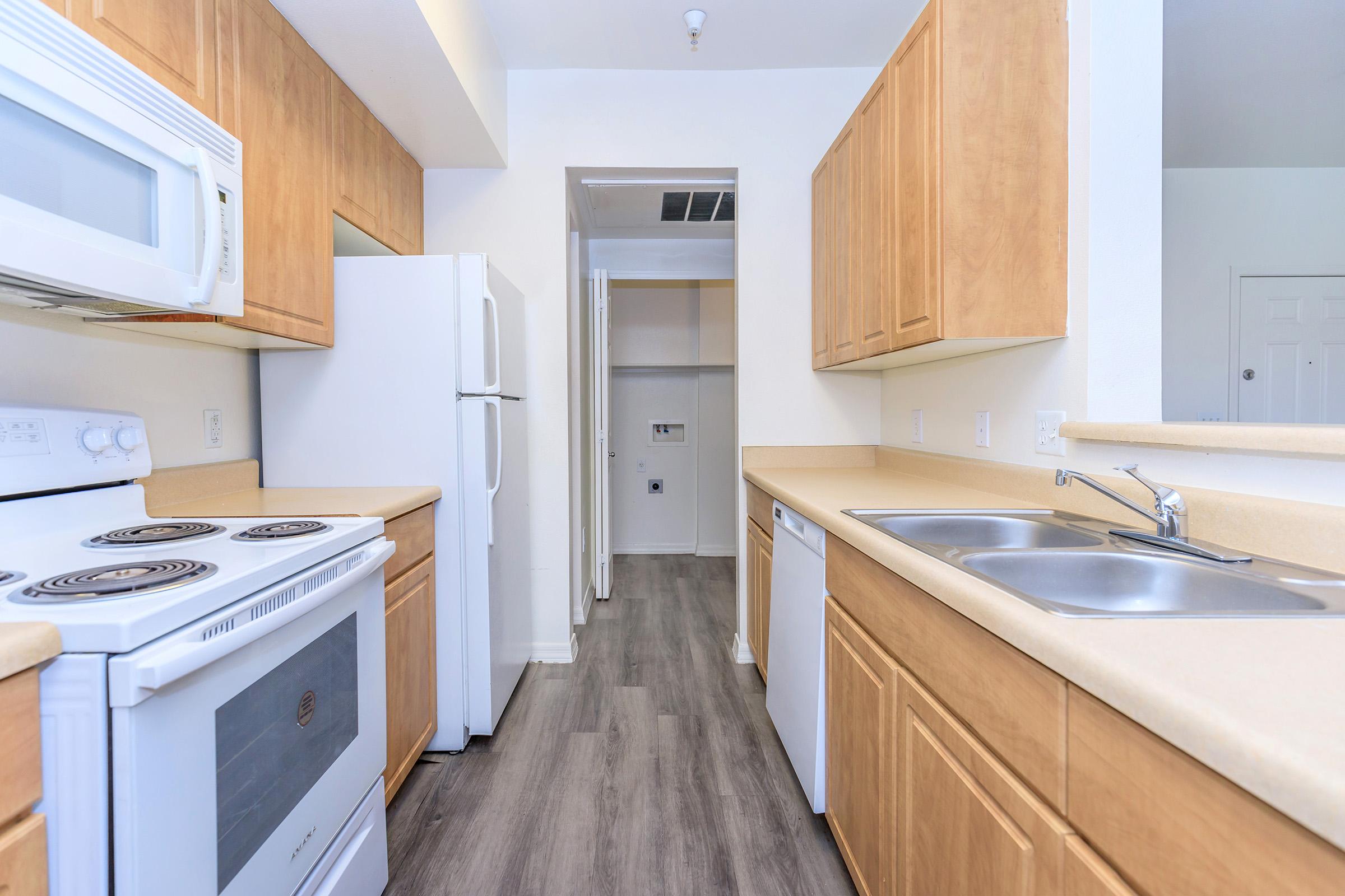 A modern kitchen featuring light wood cabinetry, a white stove, microwave, and dishwasher. There's a double sink with a faucet, and the space includes a refrigerator. The flooring is a dark laminate, creating a contrast with the cabinets. A doorway leads to another room in the background.