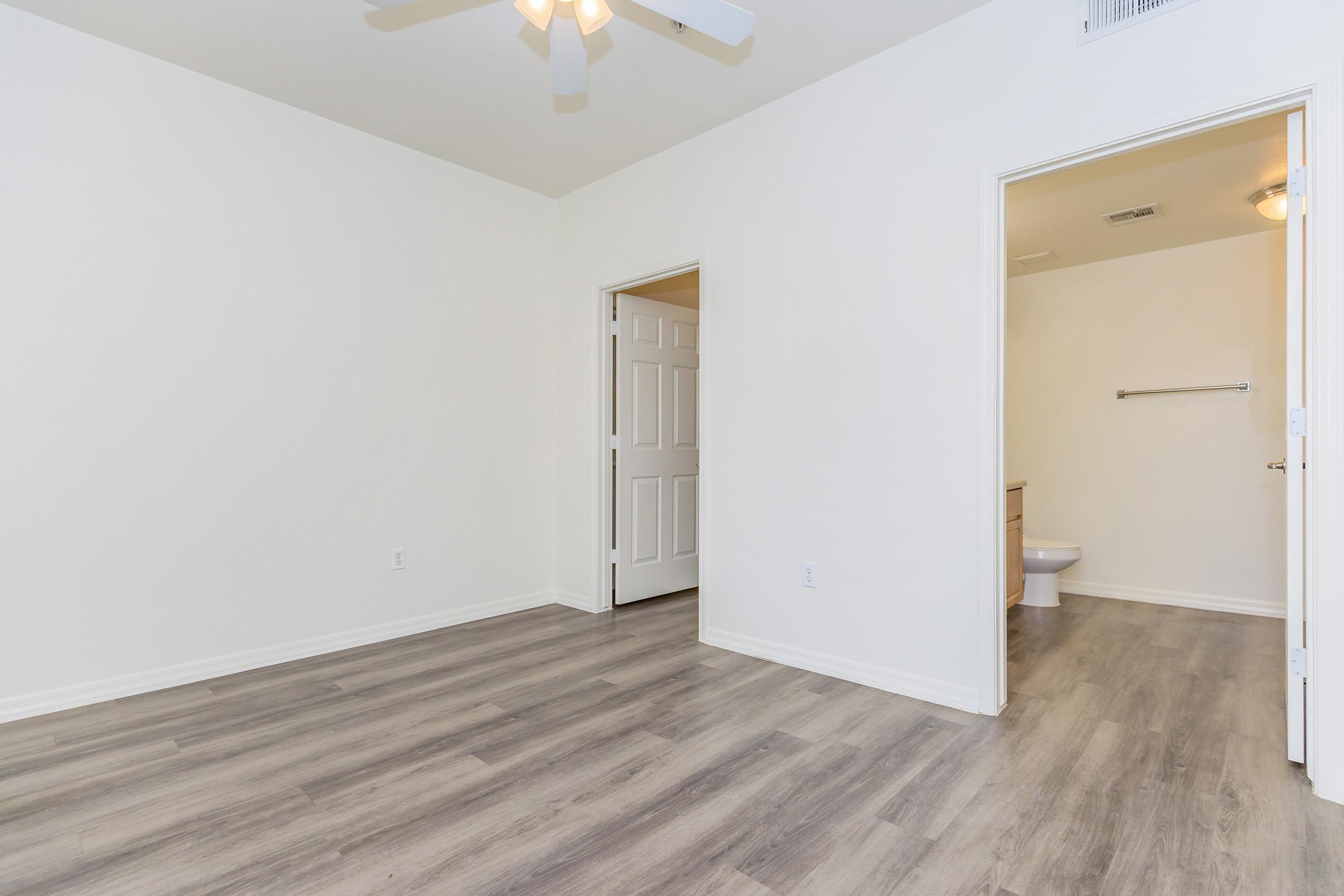 A spacious, empty room featuring light-colored walls, a ceiling fan, and new laminate flooring. There's an open doorway on one side leading to a bathroom, and another doorway leading to a hallway. The room is bright and airy, with natural light coming in.