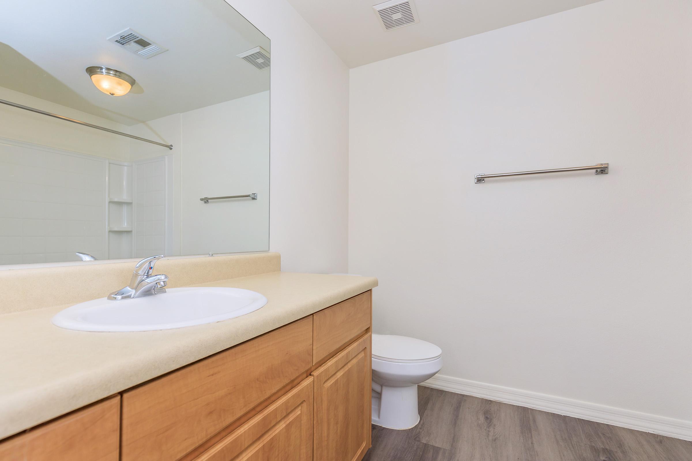 A clean and modern bathroom featuring a light-colored countertop with a sink and faucet, a large wall mirror above it, a toilet on one side, and a towel rack on the wall. The floor has a wood-like appearance, and the walls are painted light, providing a spacious feel.