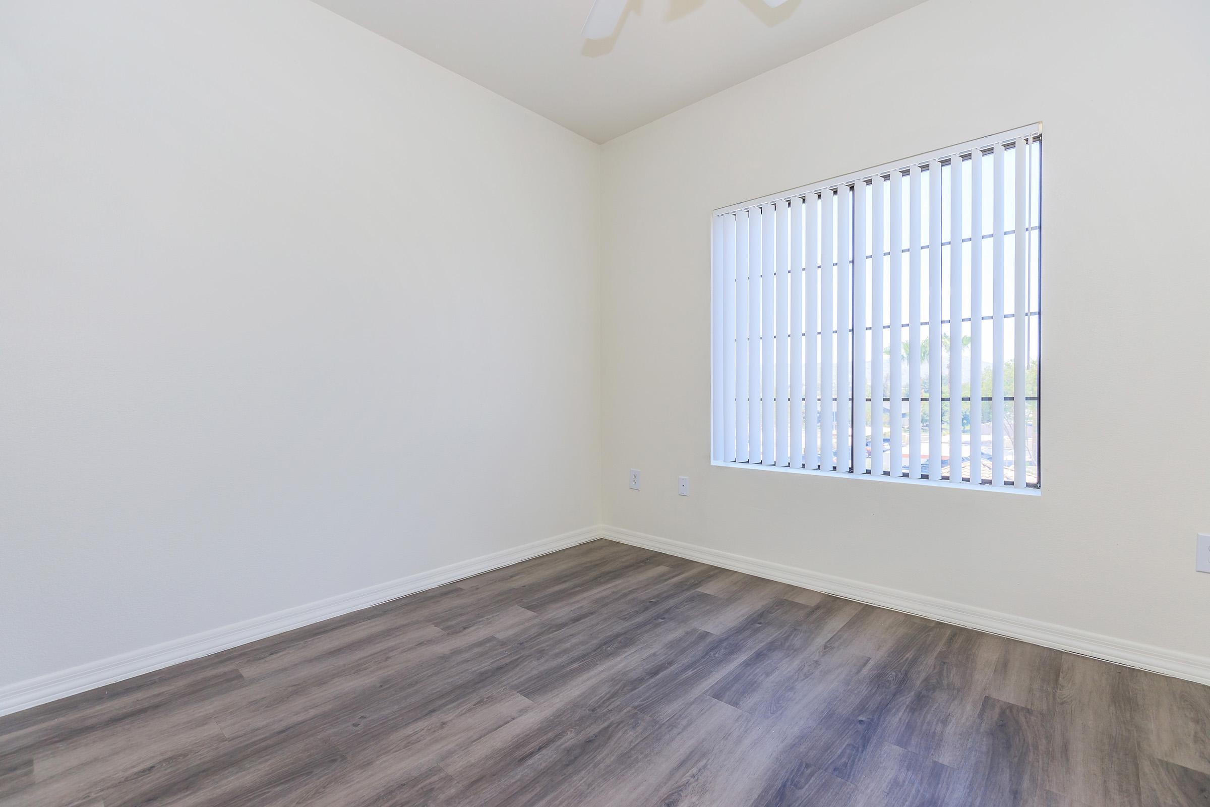 Empty room with light-colored walls and a wooden floor. A window with vertical blinds allows natural light to enter, creating a bright and airy atmosphere. The room is unfurnished and has minimal decor, emphasizing its spaciousness and simplicity.