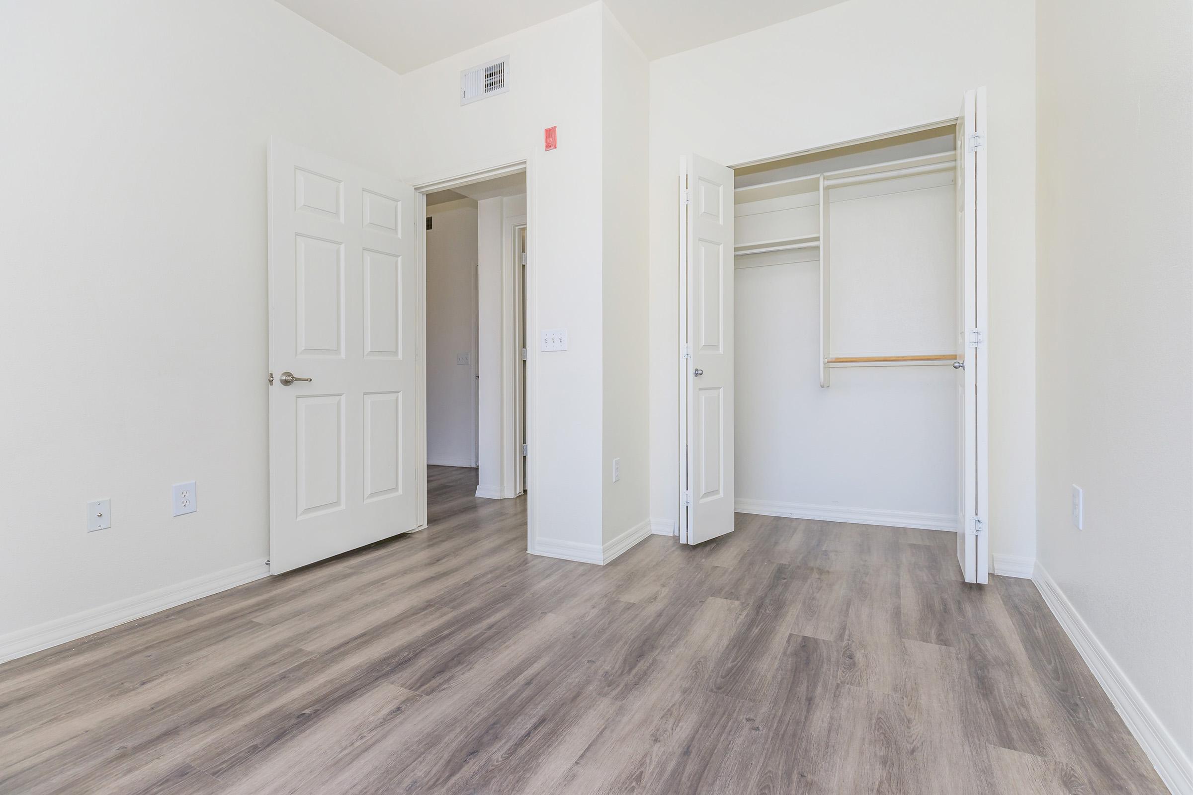 A spacious, empty room featuring light-colored walls and a wooden floor. Two doors are slightly ajar, revealing a closet on one side. Natural light highlights the room's openness and simplicity. Ideal for various furniture arrangements or as a blank canvas for personal decoration.