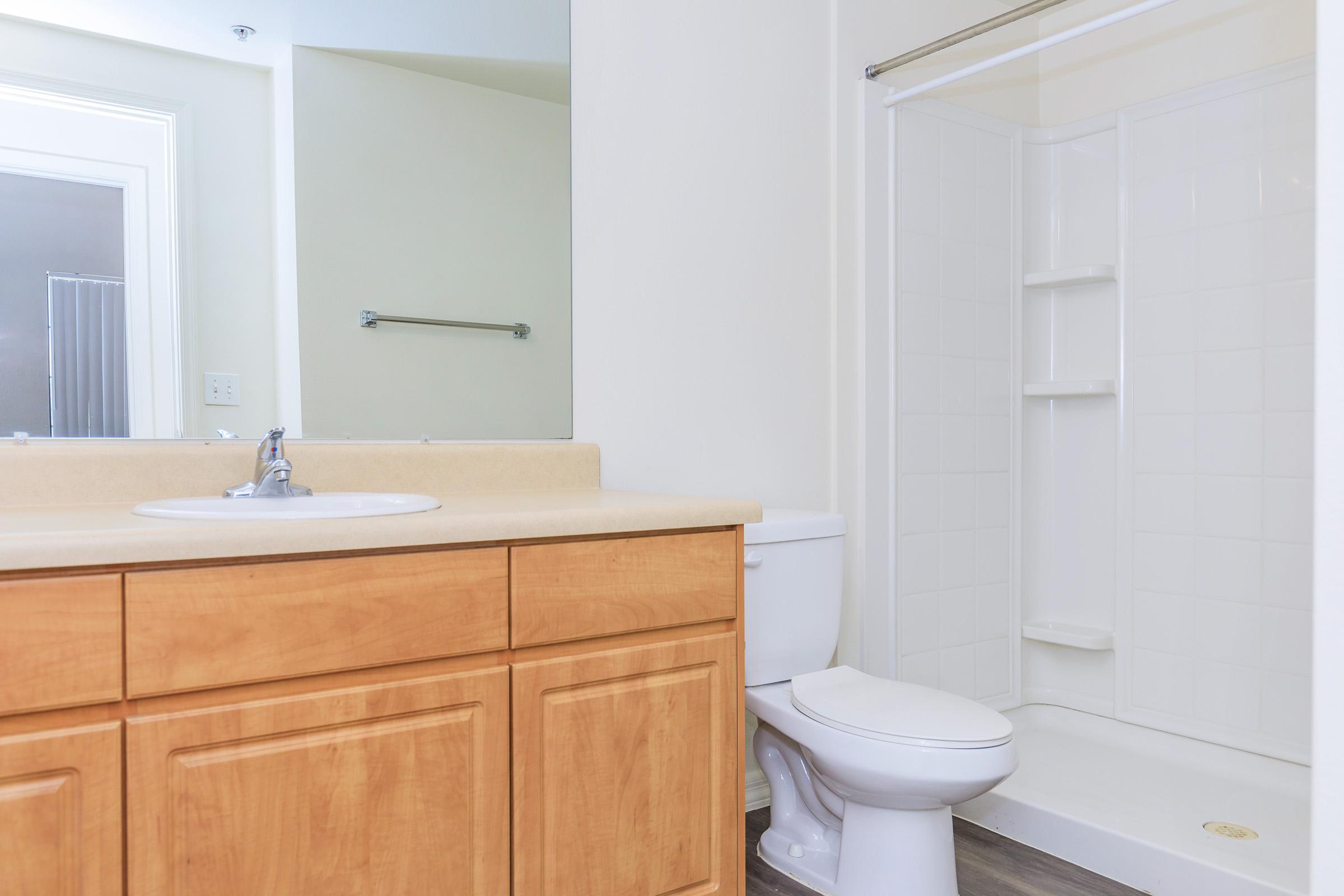 A clean and modern bathroom featuring a light-colored countertop with a sink, wooden cabinetry, a toilet, and a shower stall with white tiles. A large mirror is mounted on the wall above the sink, and the overall color scheme is neutral and bright.