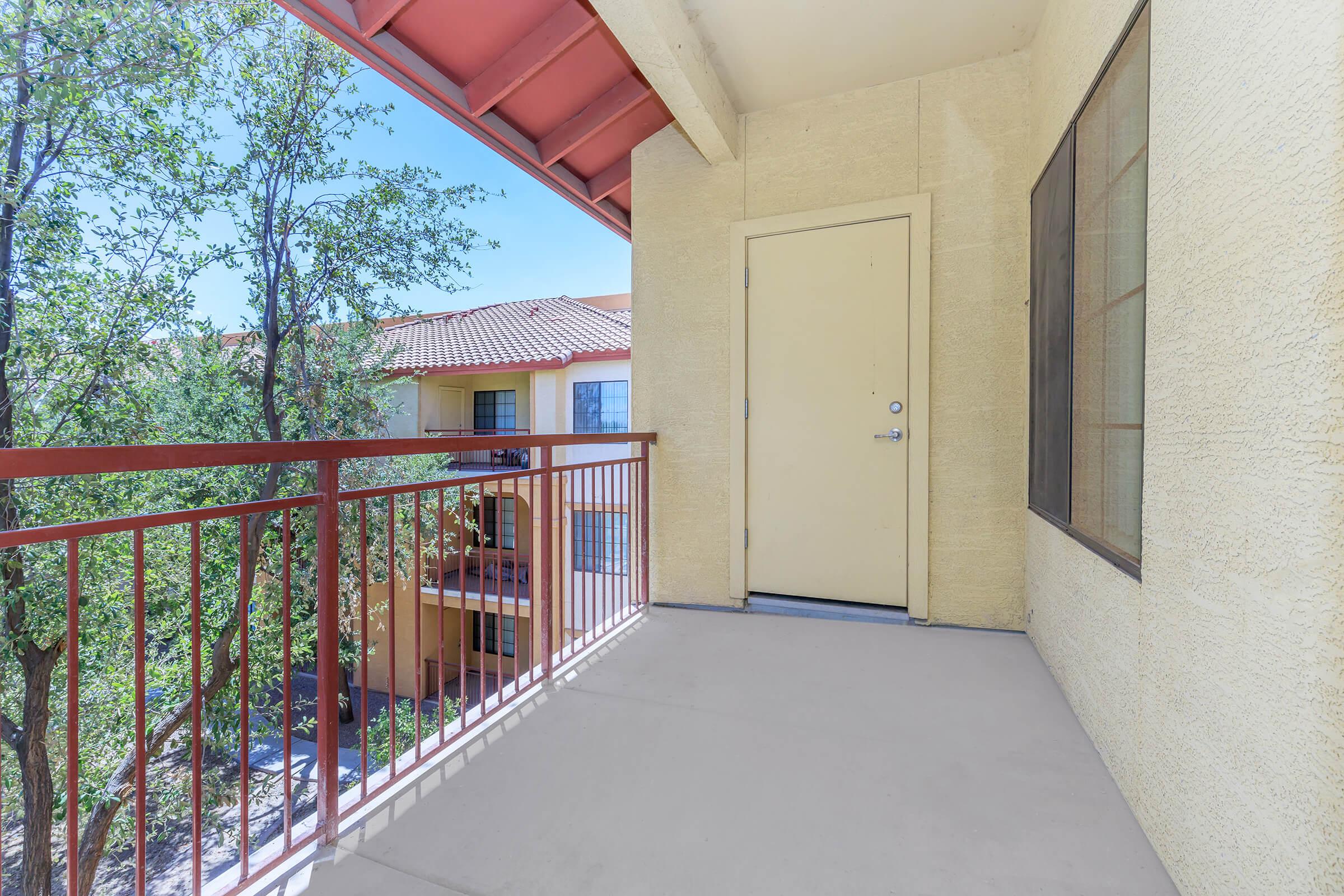 A view of a small balcony with a red railing, featuring a closed beige door and a large window. Surrounding trees and buildings are visible in the background, and the sky is clear and blue. The scene conveys a bright, inviting atmosphere.