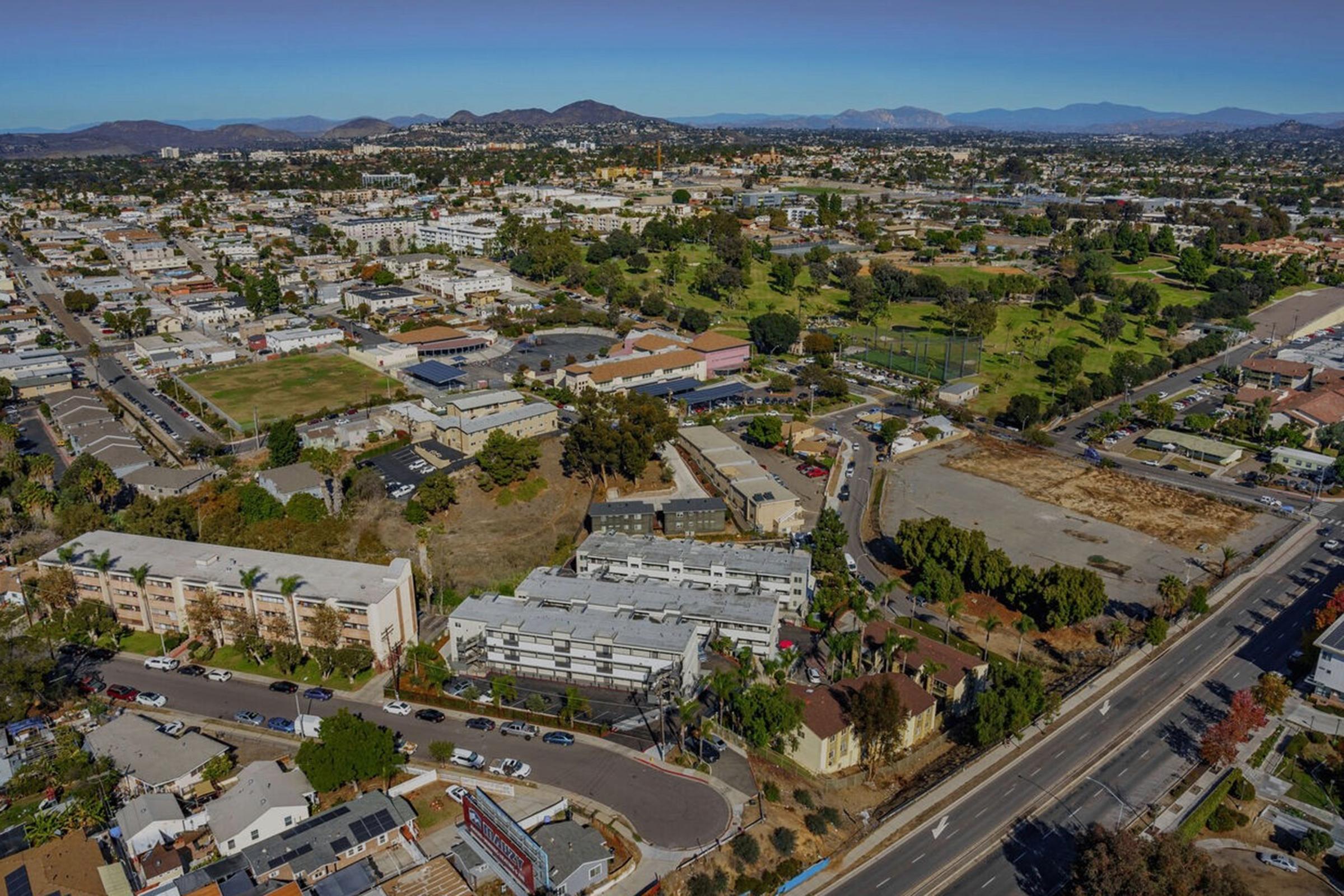 Aerial view of a suburban area featuring a mix of residential buildings, commercial properties, and green spaces. The landscape includes winding roads, parking lots, and patches of open land, with mountains visible in the background under a clear blue sky.