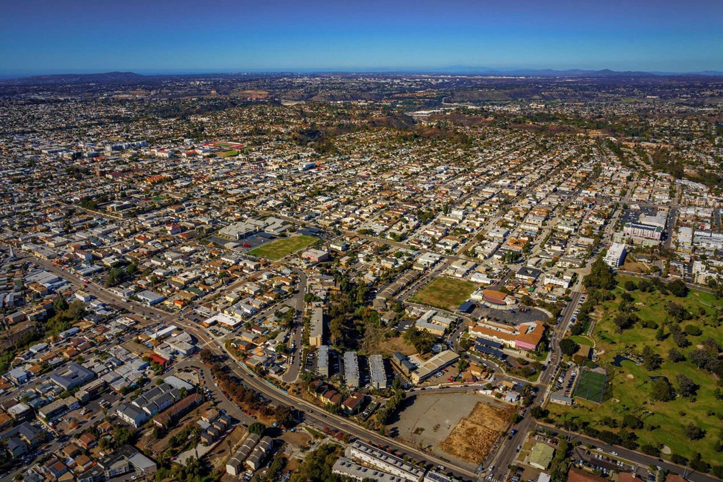 Aerial view of a densely populated urban area featuring residential neighborhoods, roads, and patches of green spaces. The landscape includes a mix of single-family homes and commercial zones, with hills in the background under a clear blue sky.