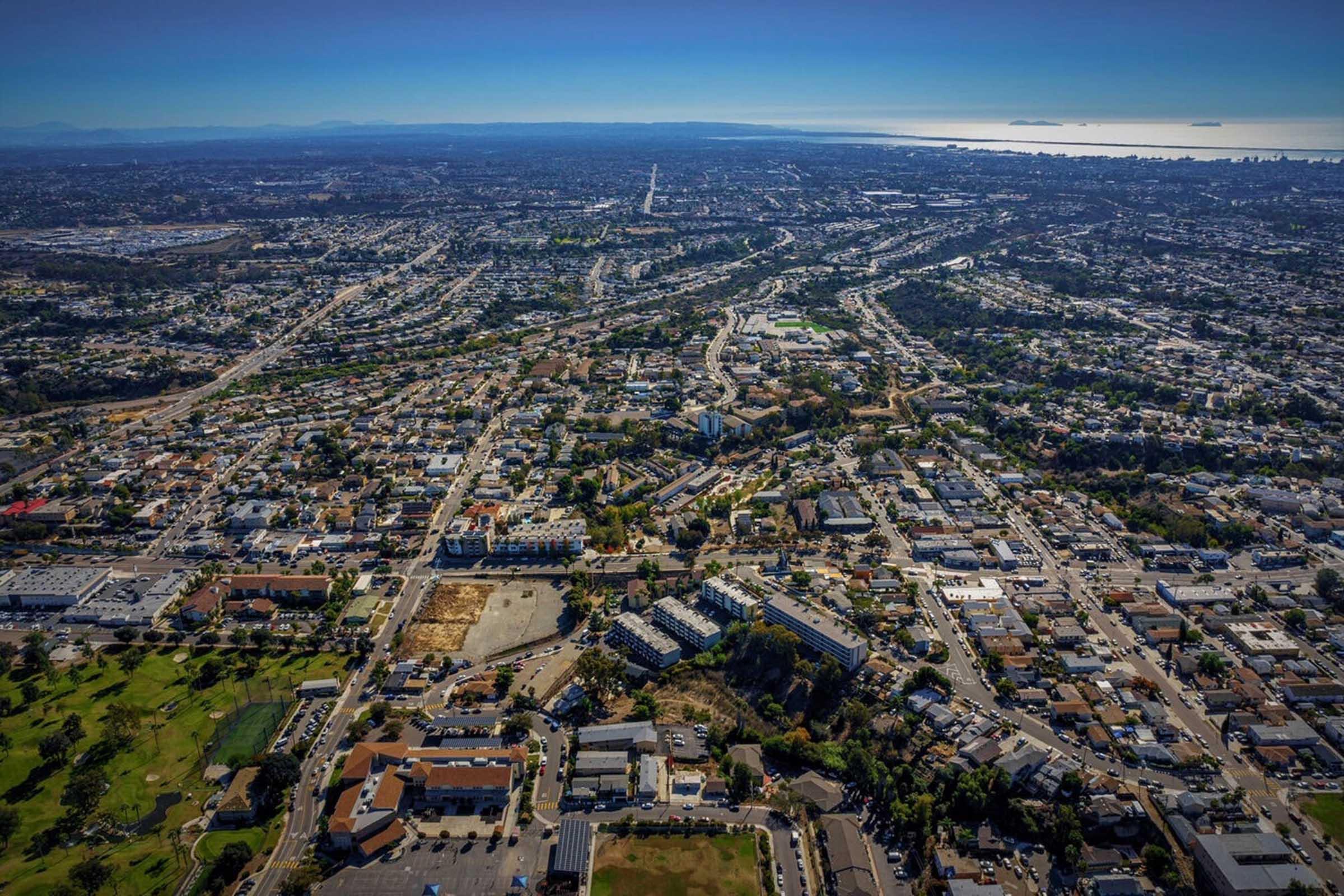 Aerial view of a sprawling urban landscape featuring a network of roads, buildings, and green spaces. In the distance, the coastline and ocean are visible, while the skyline of a city can be seen on the horizon. The scene captures the complexity and density of a developed area with both residential and commercial structures.