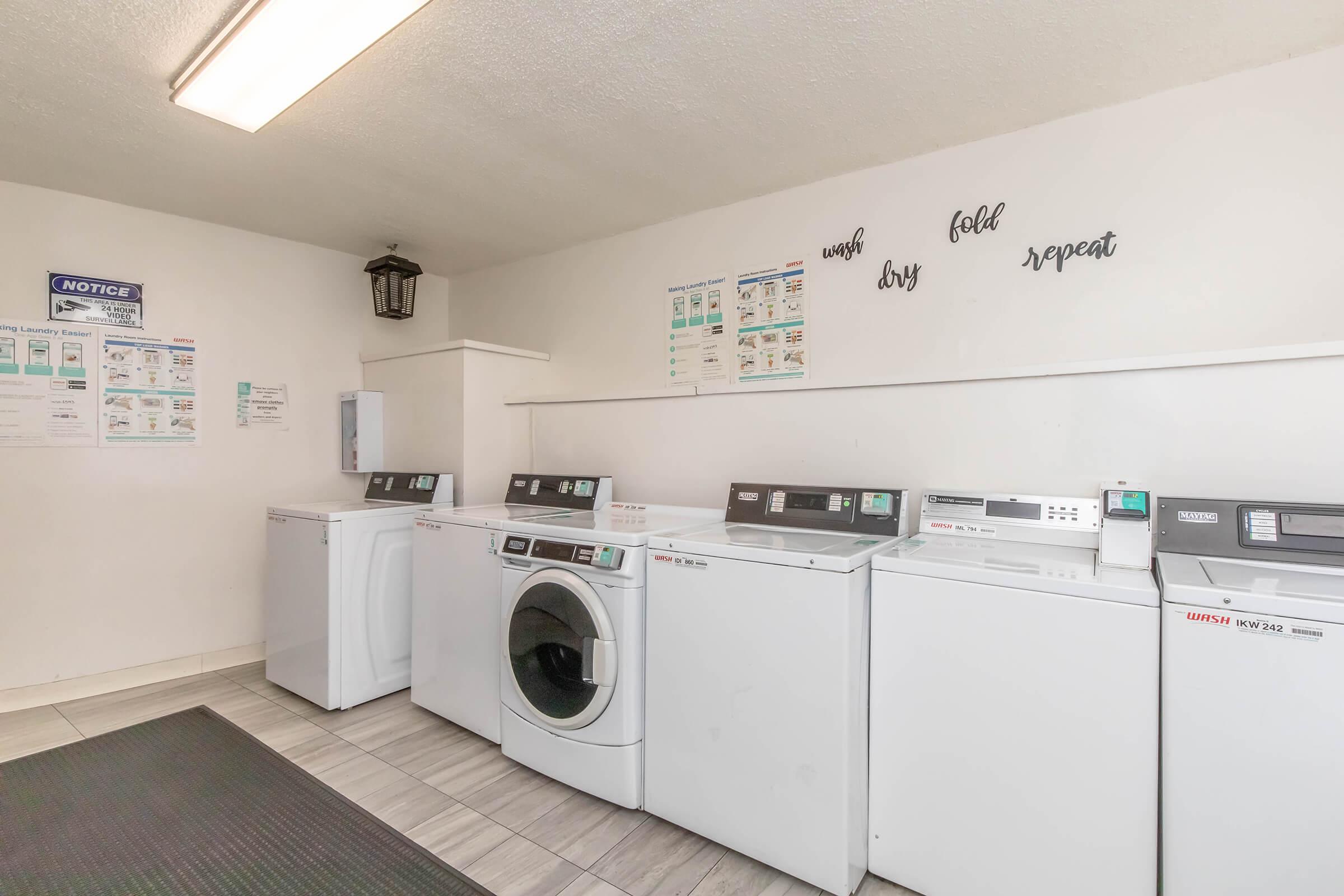 A clean and well-lit laundry room featuring several washing machines and dryers lined up against a wall. Above the machines are decorative writings that read "wash, dry, fold, repeat." The space is organized, with informational posters on the wall and a mats on the floor.