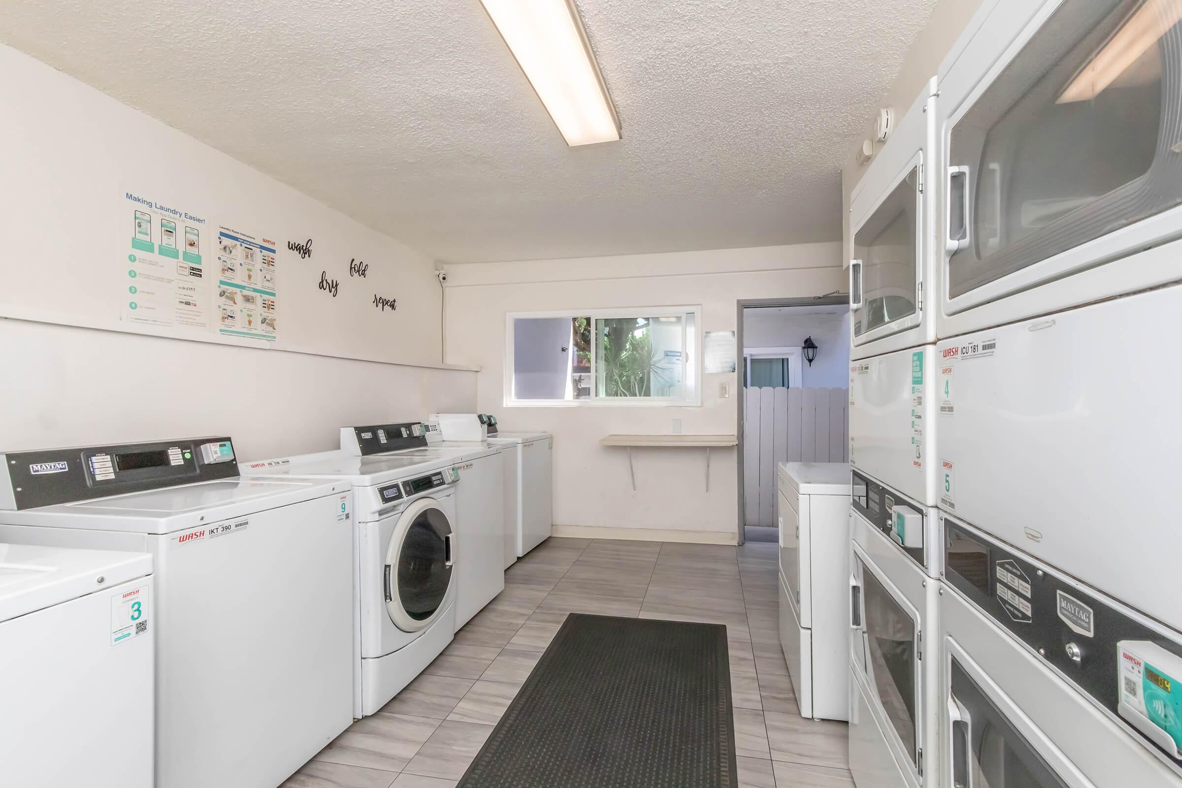 A clean, well-lit laundry room featuring multiple washing machines and dryers lined along the walls. There's a mat on the floor, a window allowing natural light, and informational posters on the wall. The space is organized and inviting for users.
