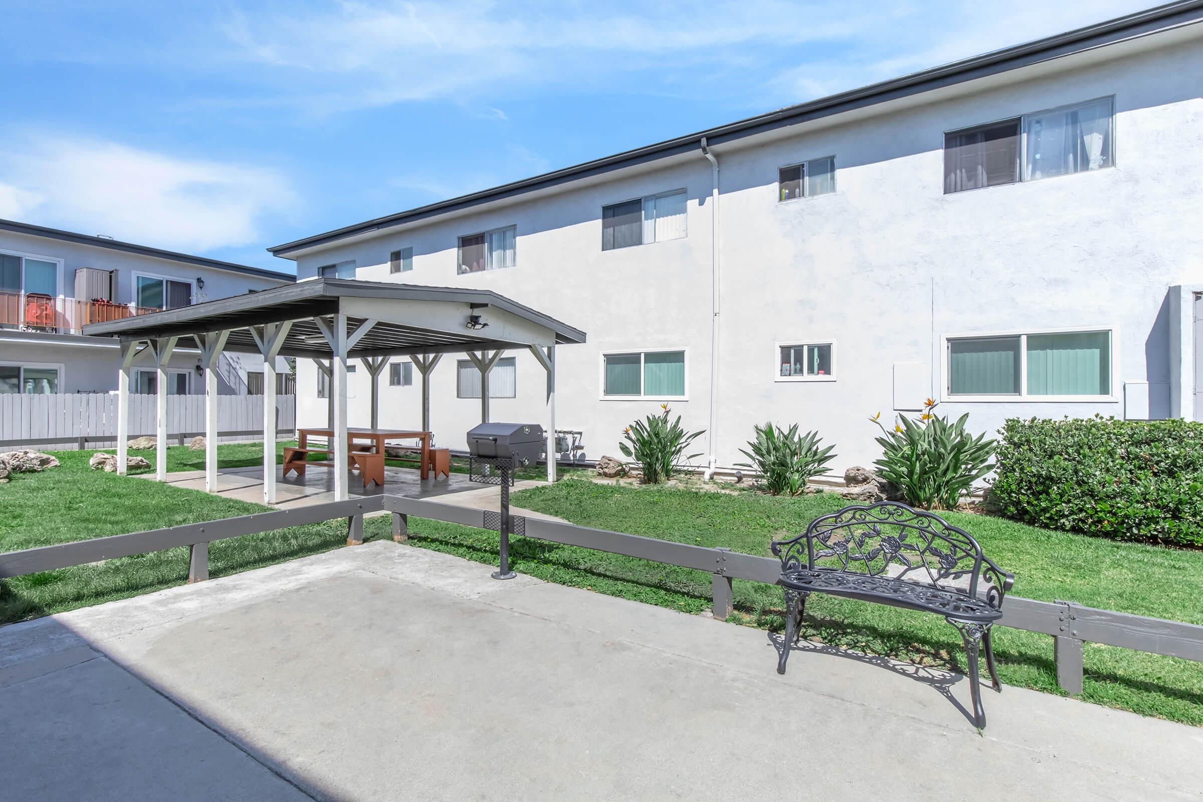 A view of a courtyard area featuring a gazebo with a grill, surrounded by well-maintained grass and shrubs. The background shows a white multi-unit residential building with several windows. There is a bench facing the gazebo, creating a cozy outdoor space.