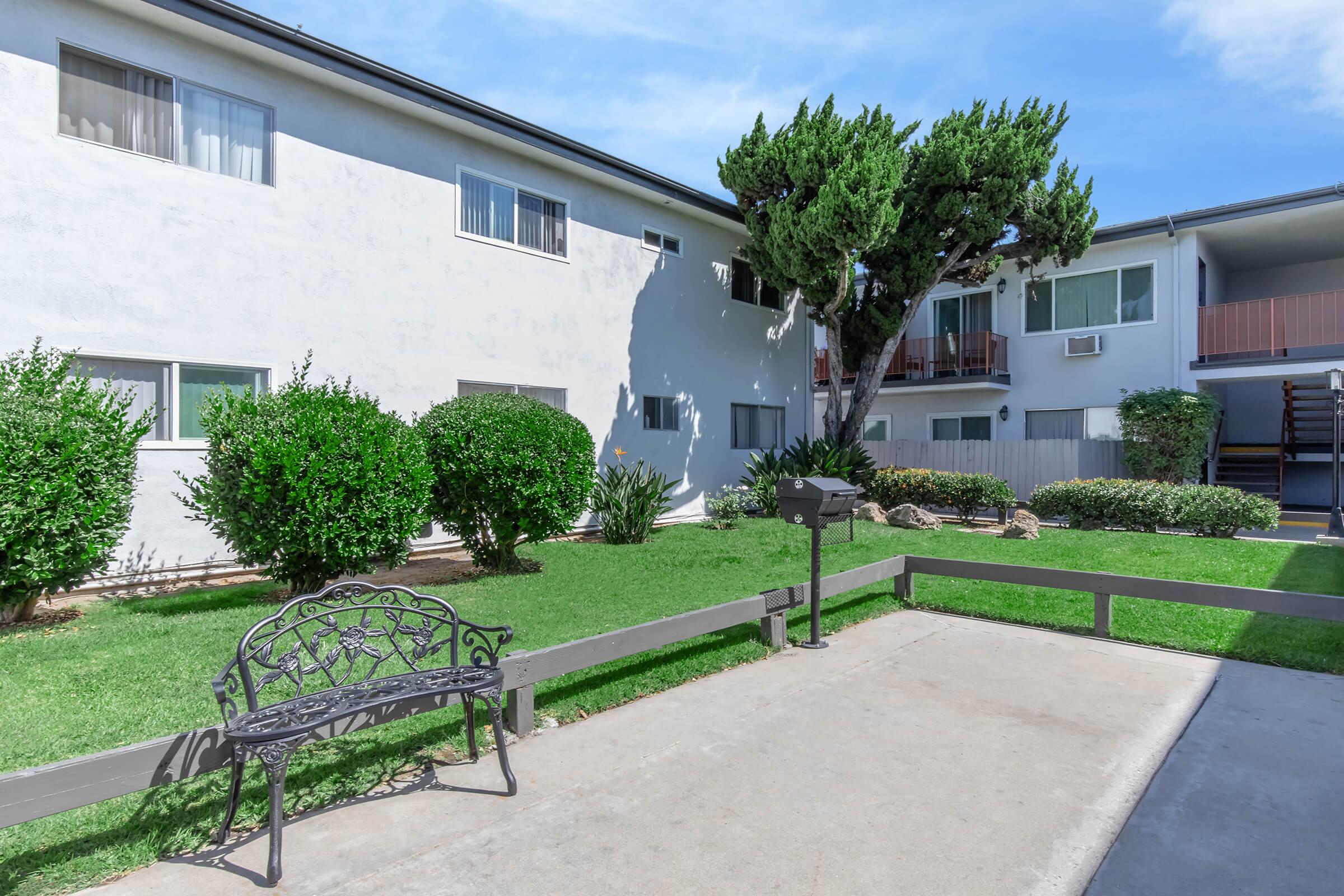 A well-maintained courtyard with lush green grass and neatly trimmed bushes, featuring a black wrought-iron bench. In the background, there are two multi-story residential buildings with balconies and windows, set under a clear blue sky. A shaded area near the bench adds to the inviting atmosphere.