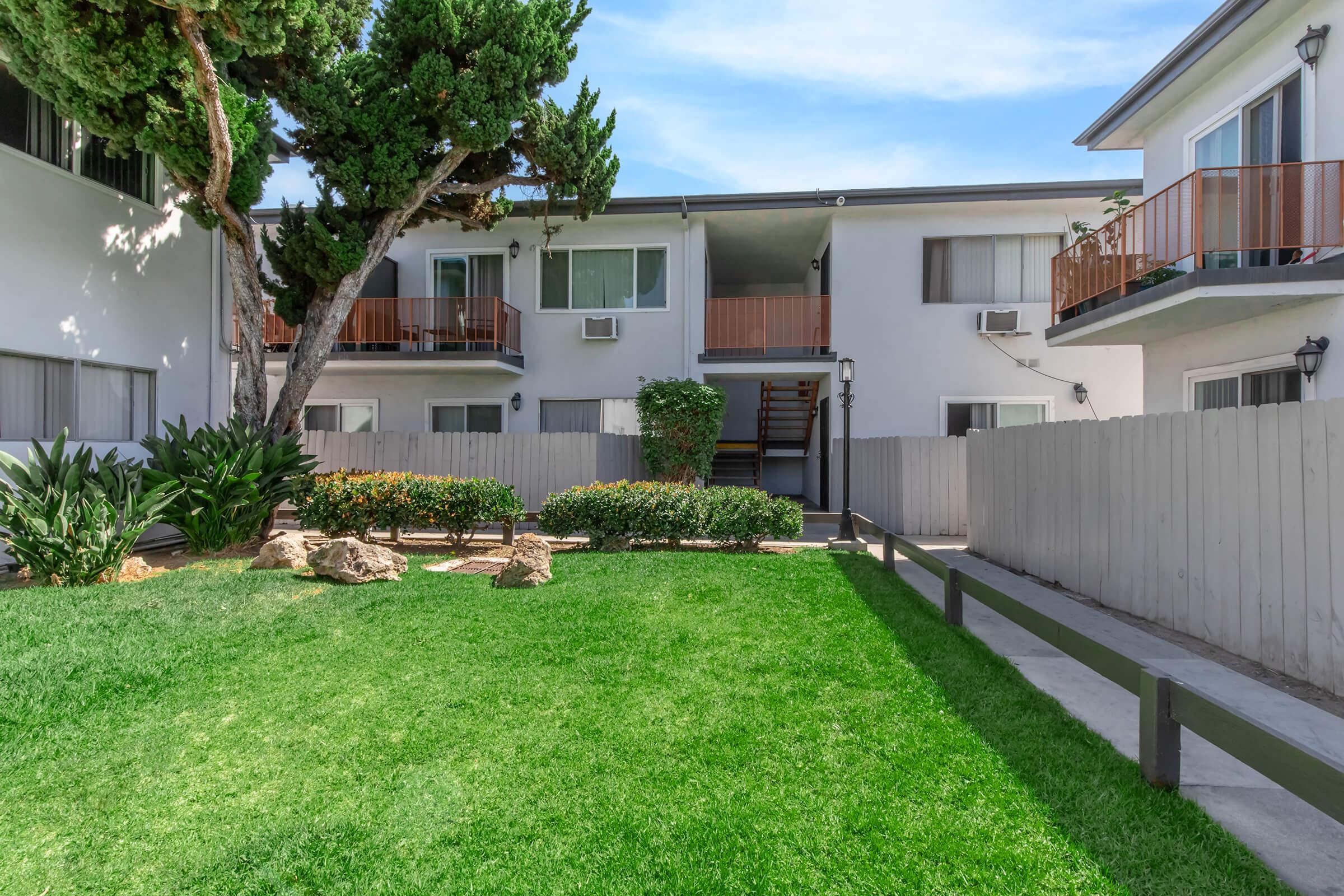 A grassy area with well-maintained landscaping featuring shrubs and rocks, surrounded by buildings with balconies. A pathway leads to the entrance of the apartments, and there are trees providing shade. The scene is bright and inviting, reflecting a quiet residential atmosphere.