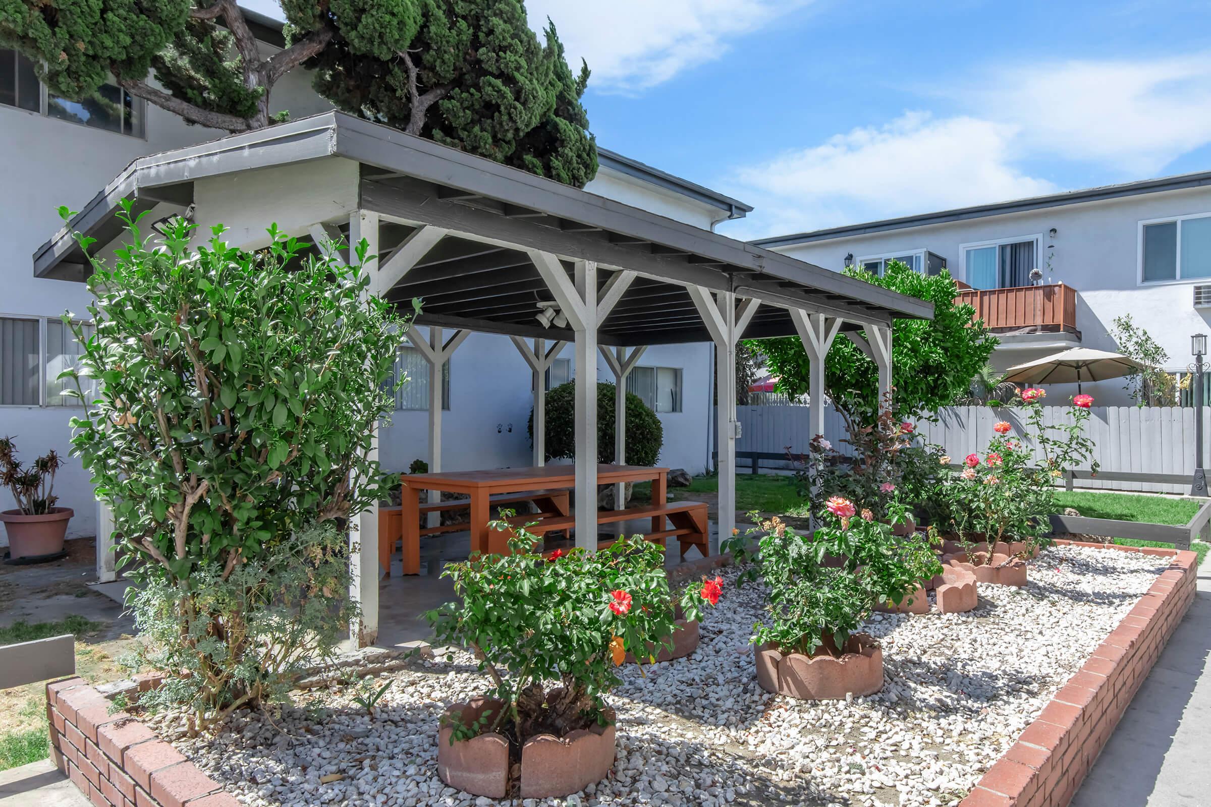 A wooden gazebo surrounded by flowering plants and shrubs, with a gravel base. The gazebo features a large wooden table and benches, set in a well-maintained garden area. In the background, there are residential buildings and a blue sky with a few clouds.
