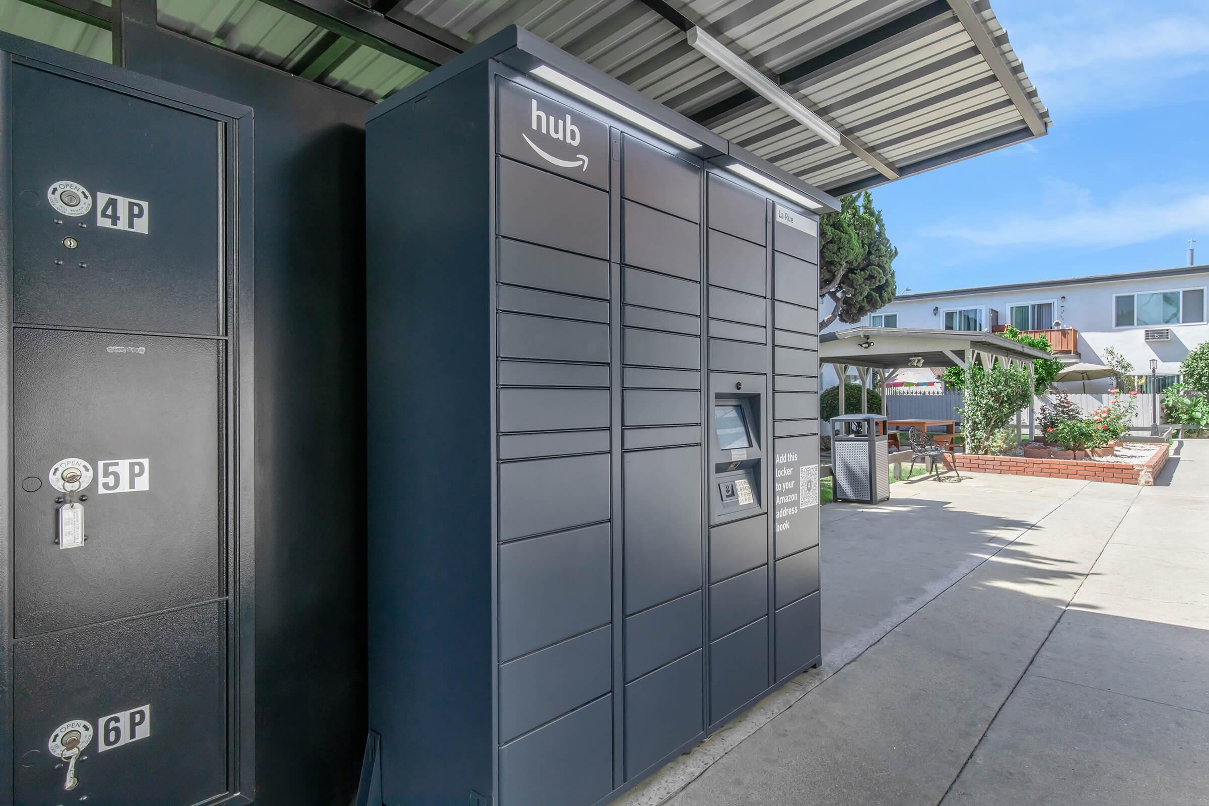 A row of Amazon Hub lockers in a shaded outdoor area, featuring several compartments for package pickup. Nearby, there are residential buildings and a landscaped courtyard, creating a pleasant environment for retrieving packages.