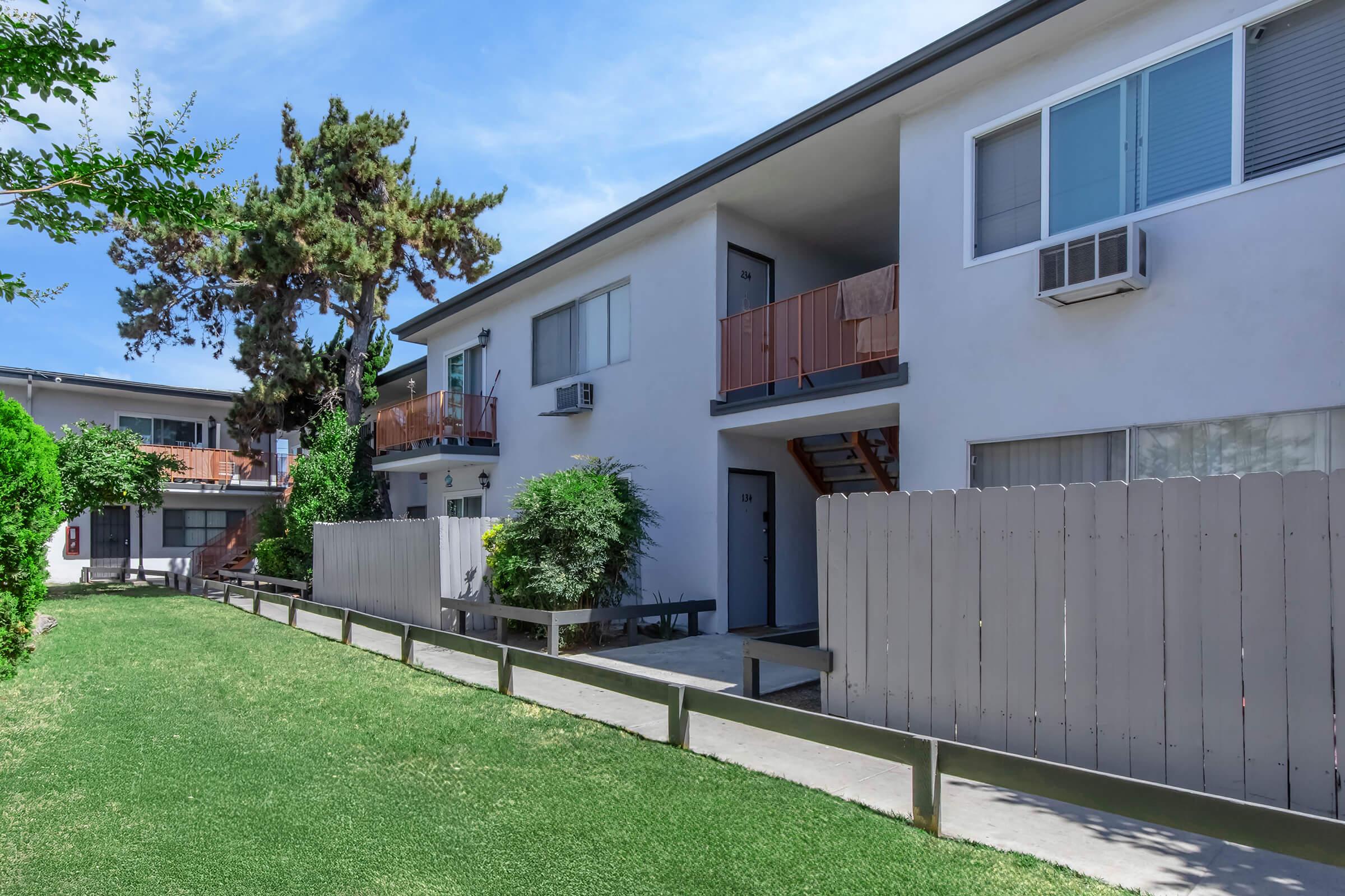 Two-story residential buildings with balconies, surrounded by well-maintained green grass and shrubs. A wooden fence runs along the pathway, leading to the entrance of the units. Air conditioning units are visible on the walls, and there are stairs leading to the upper floors. Clear blue sky overhead.