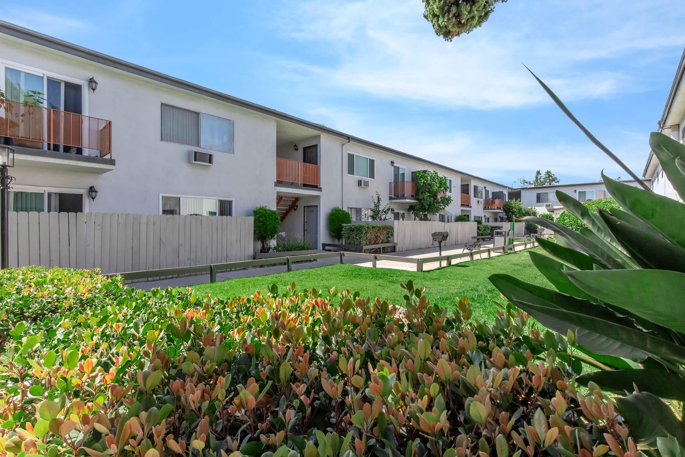 A residential complex featuring two-story buildings with balconies. Lush greenery and landscaped areas surround the pathways. The sky is clear and blue, adding to the inviting atmosphere of the community.