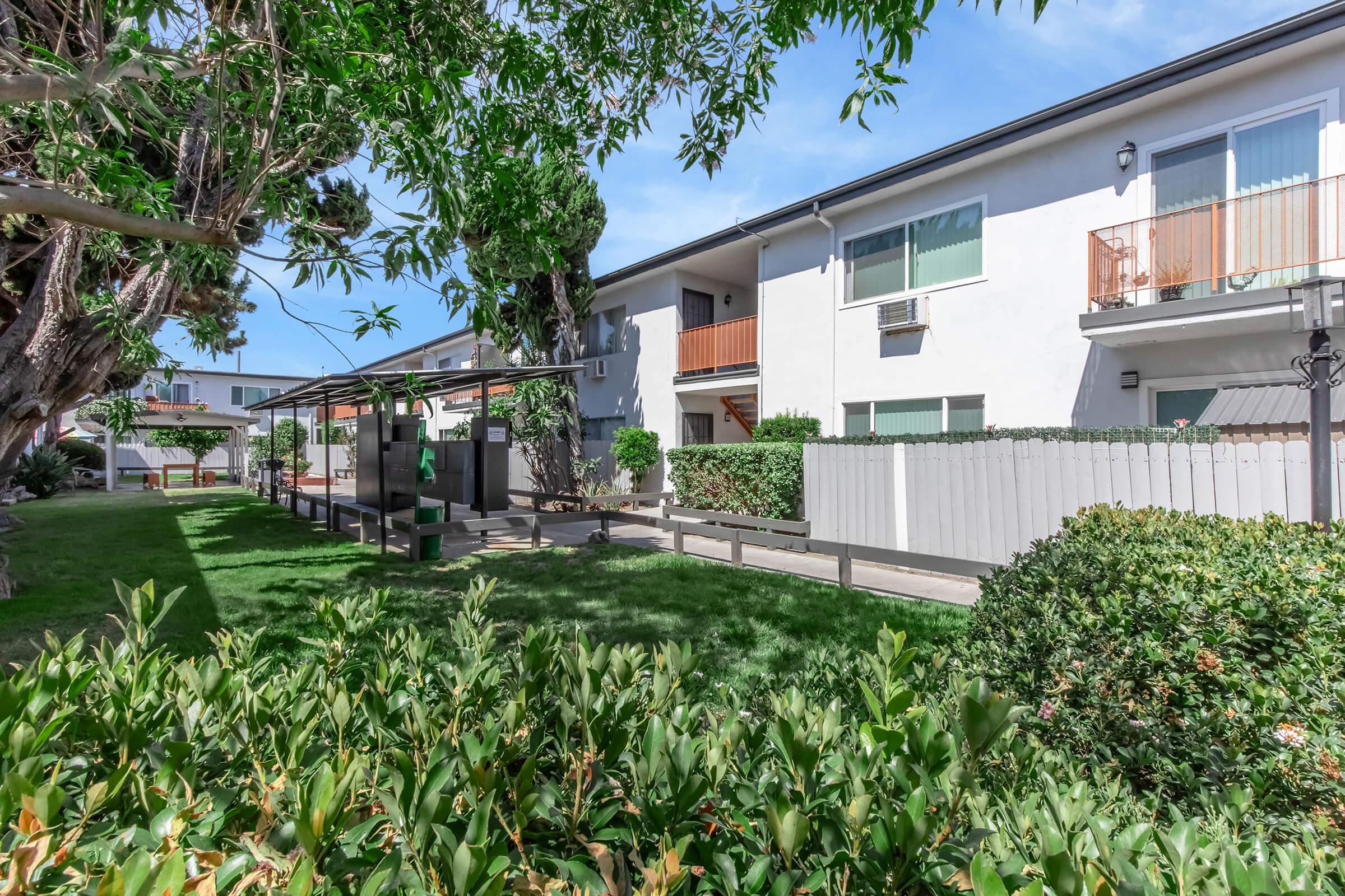 A view of a well-maintained outdoor area with green grass, shrubs, and trees. In the background, there are two-story residential buildings with balconies. An outdoor seating area is visible, along with a barbecue grill. The sky is clear, creating a bright and inviting atmosphere.