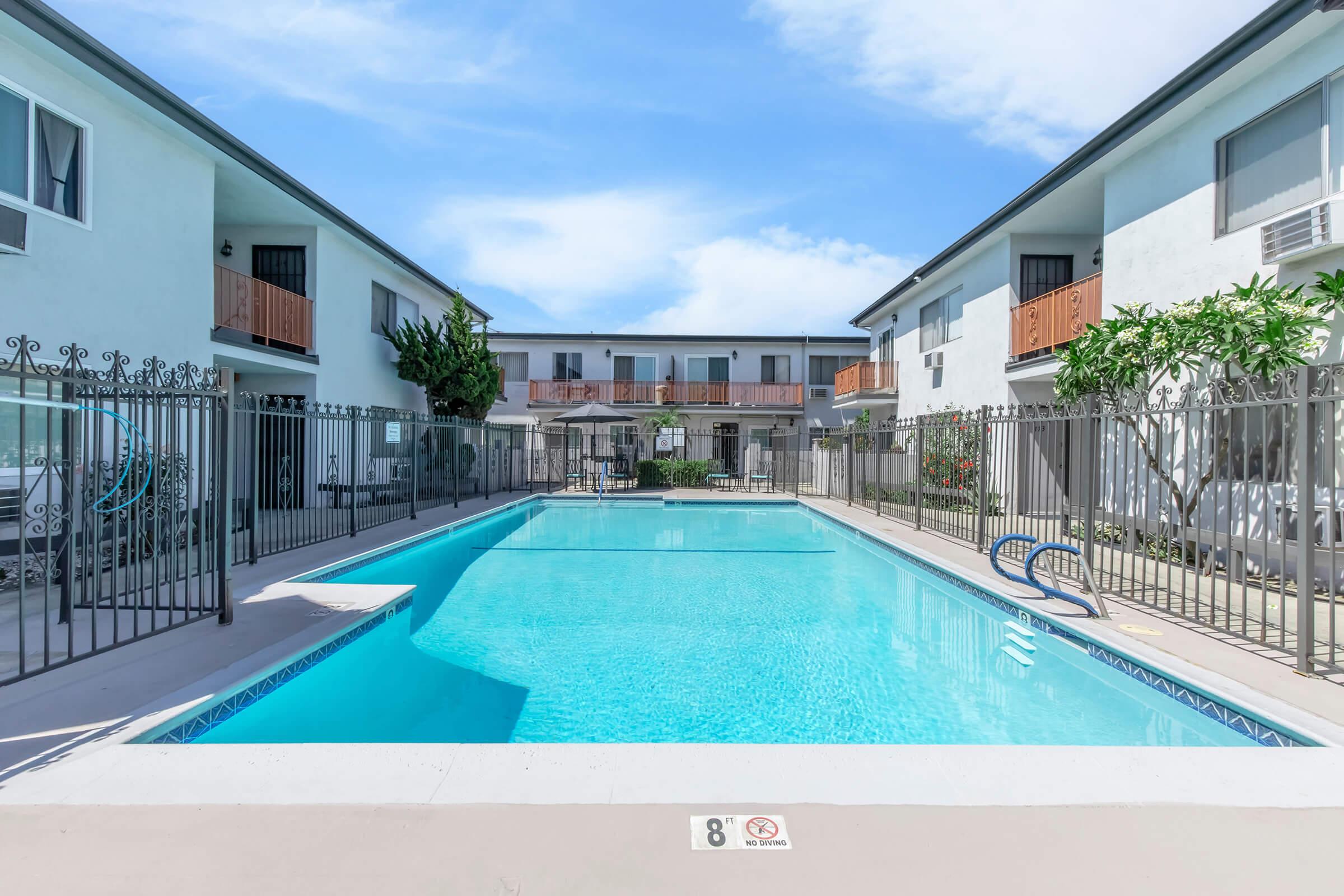 A clear swimming pool surrounded by fenced areas and two-story buildings. The pool is empty, with lounge chairs visible on the sides, and greenery along the perimeter. The sky is bright with a few clouds, indicating a sunny day.