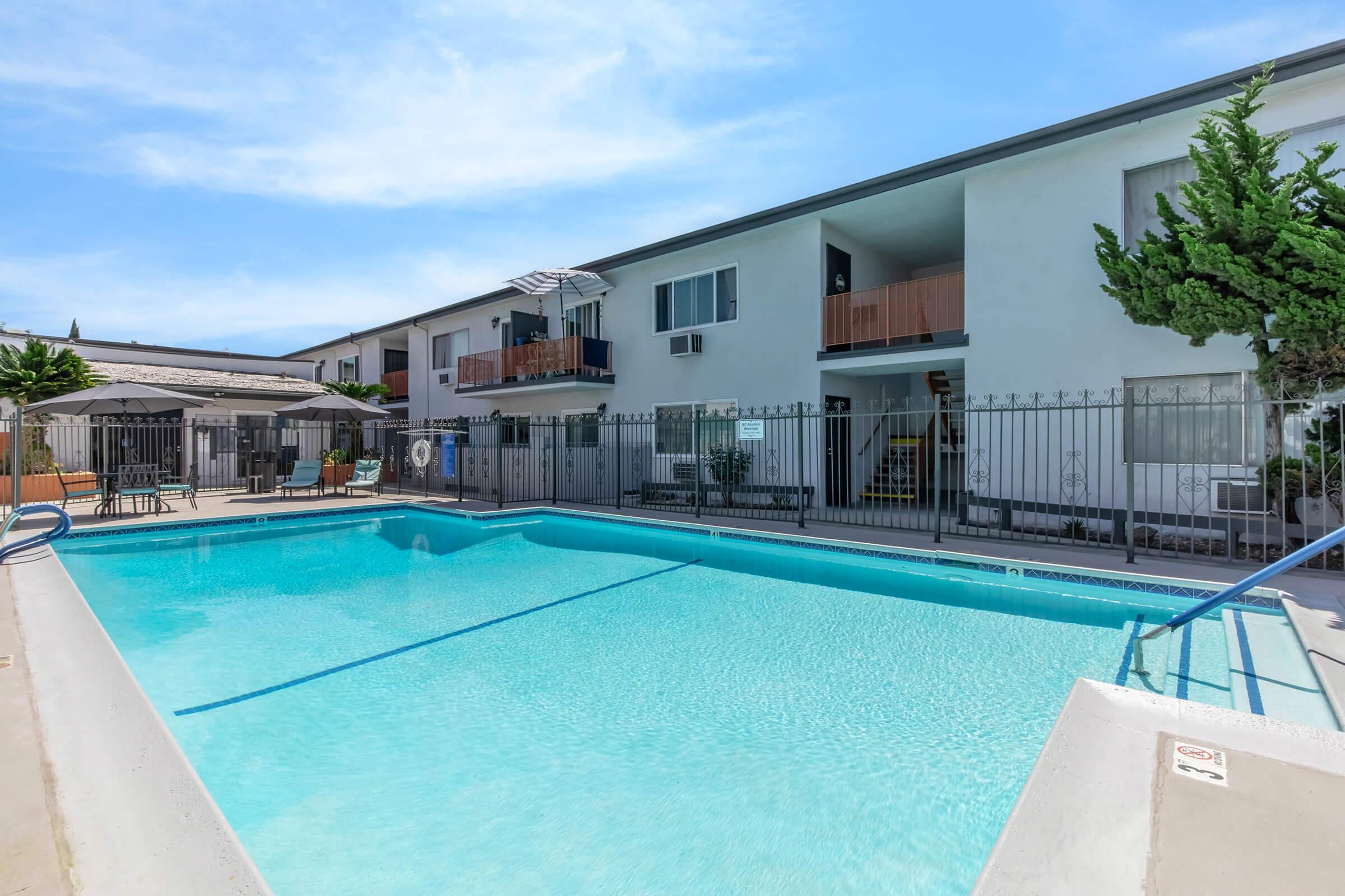 A swimming pool surrounded by a gated area, with two-story apartment buildings in the background. The pool features a clear blue water surface and stainless steel handrails. There are balconies on the second floor of the apartments and landscaping including small trees and plants around the pool area.