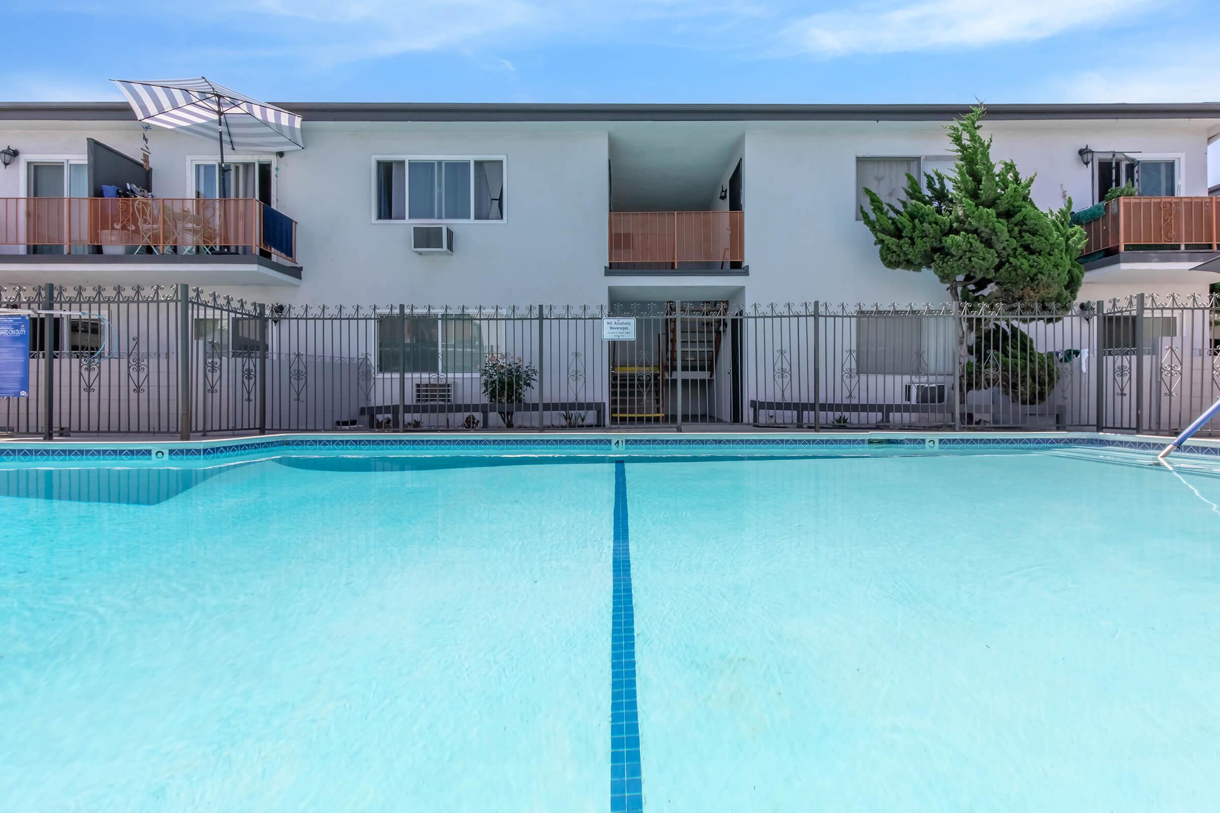 A clear swimming pool in the foreground with a neat blue tile border, reflecting the sky. In the background, a two-story building with balconies, surrounded by a fence. Lush green trees are nearby, and there's an awning over one balcony, indicating a sunny day.