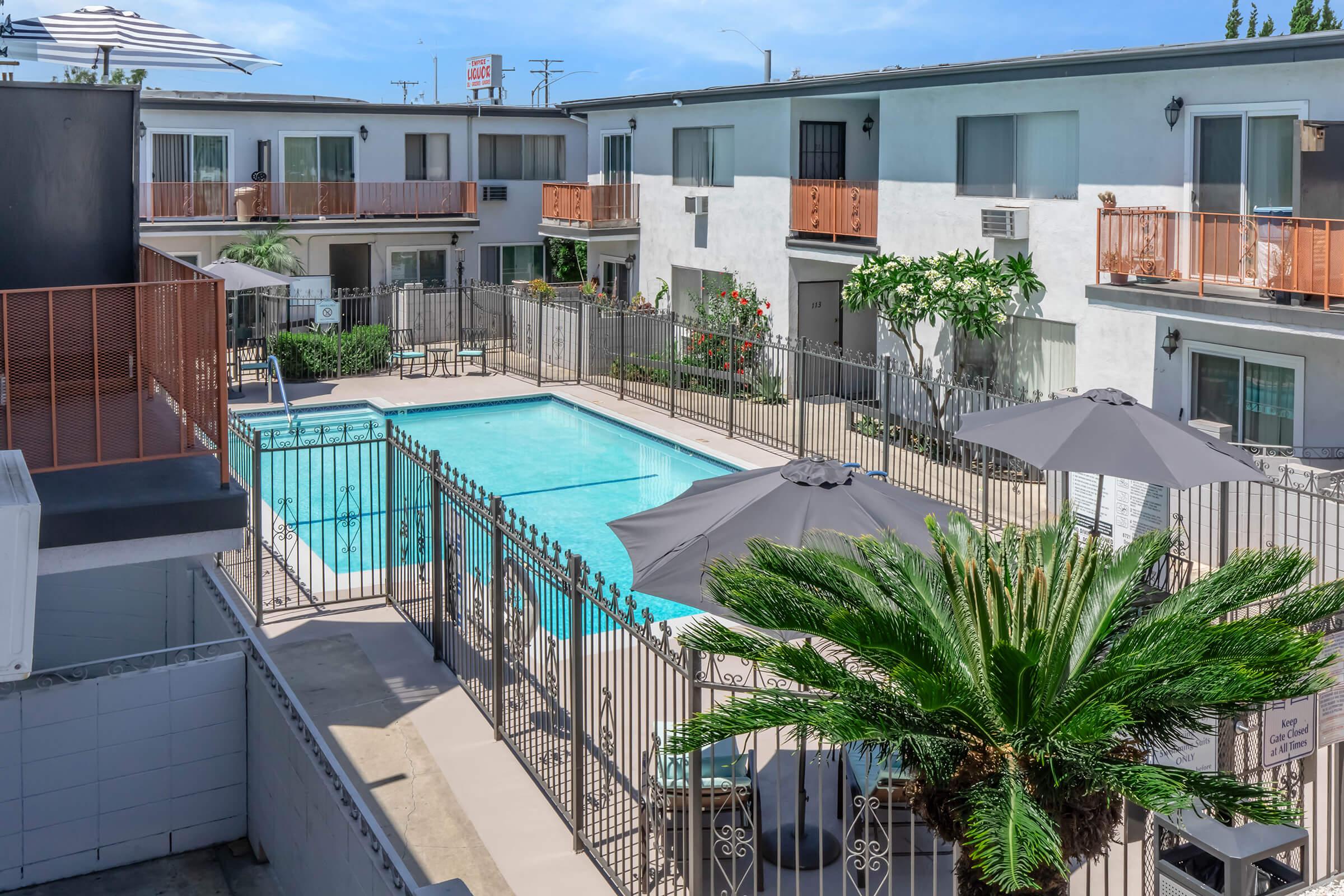 A view of a courtyard with a swimming pool surrounded by a gated fence. Several lounge chairs and large umbrellas are placed around the pool area. Nearby, there are two-story buildings with balconies, greenery, and a clear blue sky in the background.