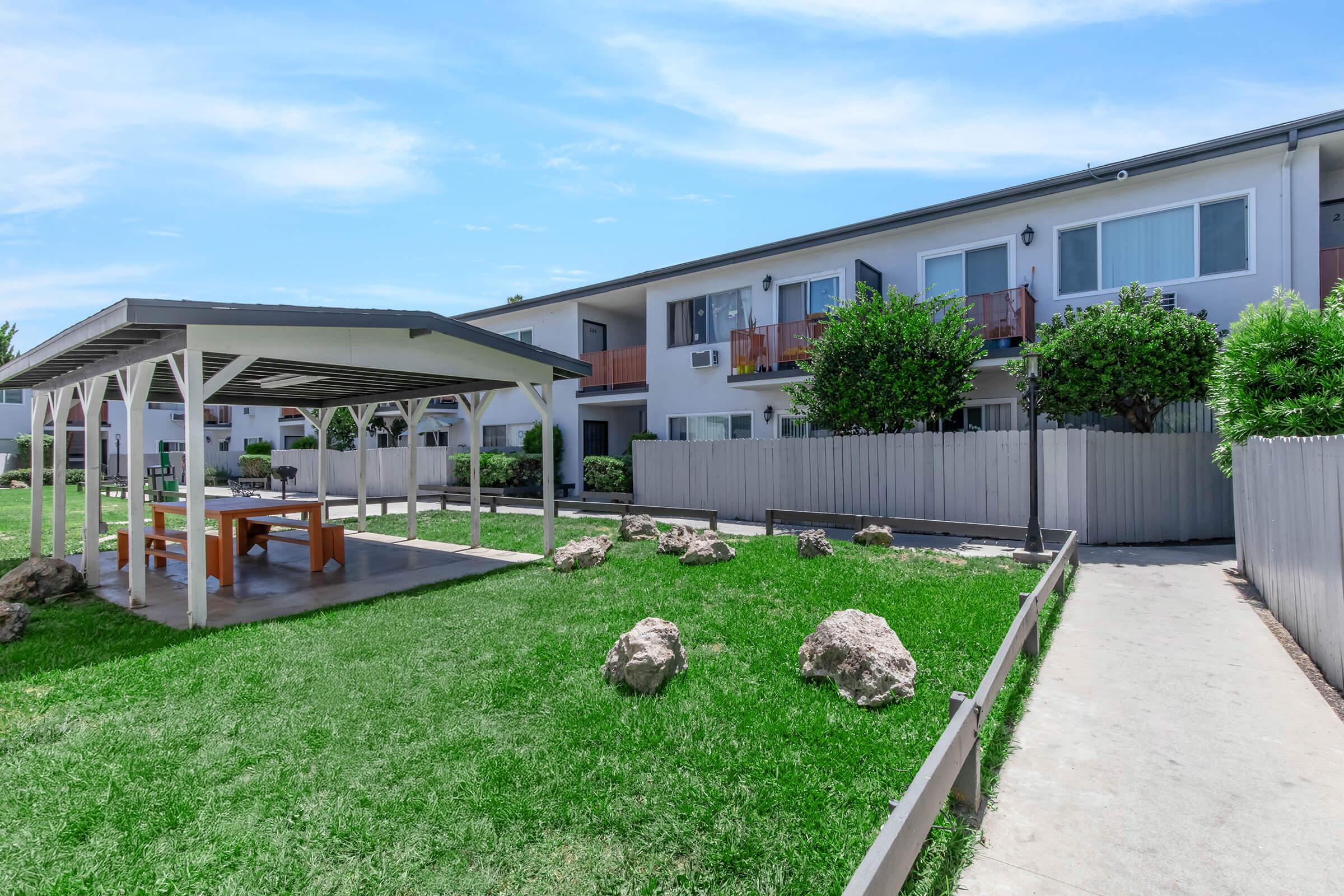 A well-maintained outdoor area featuring a gazebo with a picnic table, surrounded by green grass and decorative rocks. In the background, there are two-story apartment buildings with balconies, and a pathway leading through the space, under a clear blue sky.