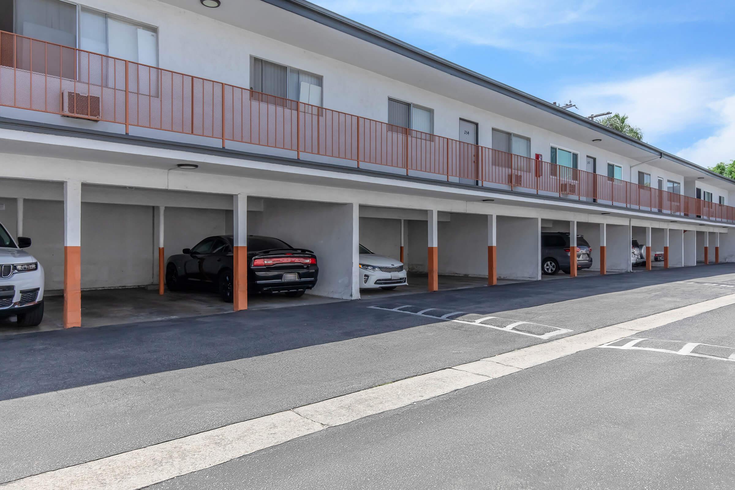 A view of a multi-unit building with a covered parking area. Several cars are parked underneath the building, while the upper level features multiple balconies. The setting is sunny with a clear blue sky and well-maintained surrounding area.